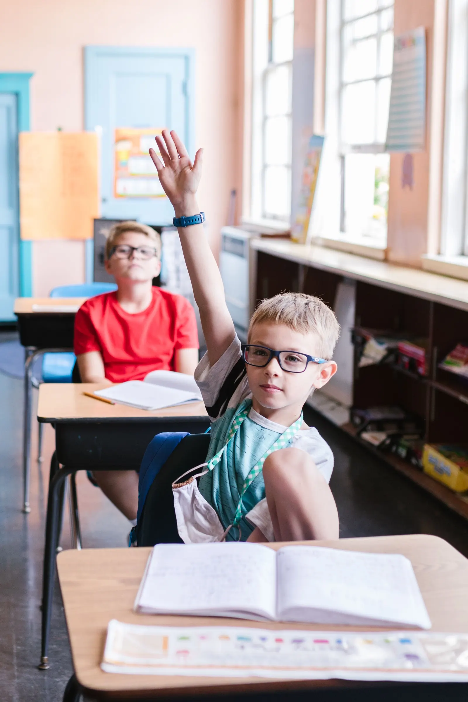 A boy in glasses raises his hand at his desk in a sunlit classroom