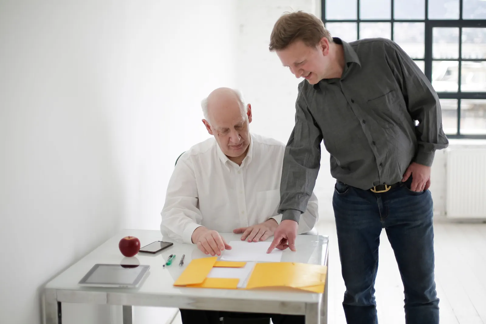 An older man at a desk and a younger man standing beside him look over papers from a yellow folder