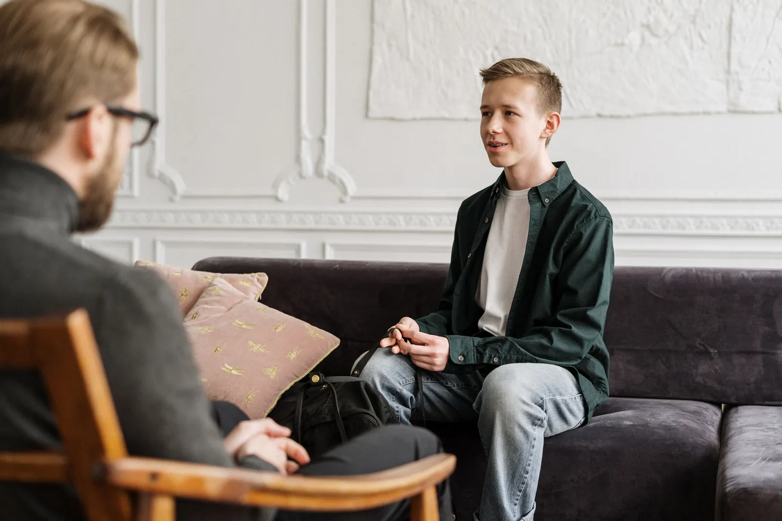 A bearded man in glasses, seen from behind, talks with a teenage boy sitting on a sofa across from him