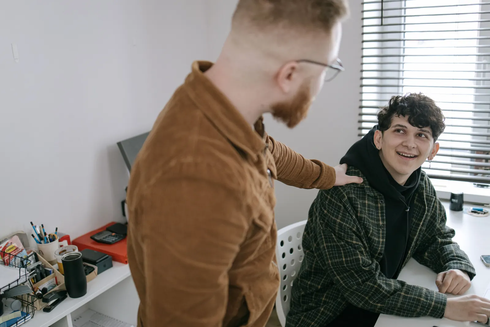 A bearded man rests a hand on a smiling teenage boy's shoulder at a desk by the window