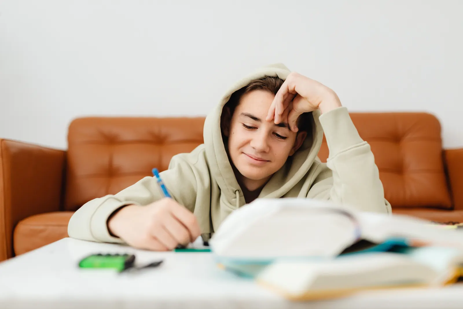 A teenage boy in a beige hoodie smiles as he writes notes, a pile of open books in front of him