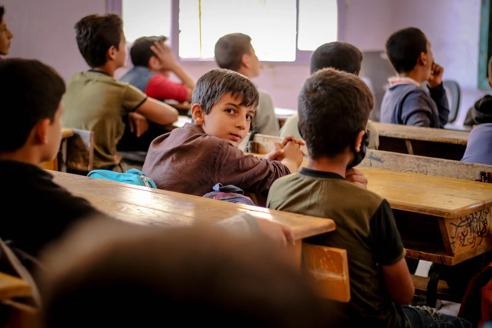 Boys at worn wooden double desks in a sunlit classroom, one turning around to look back