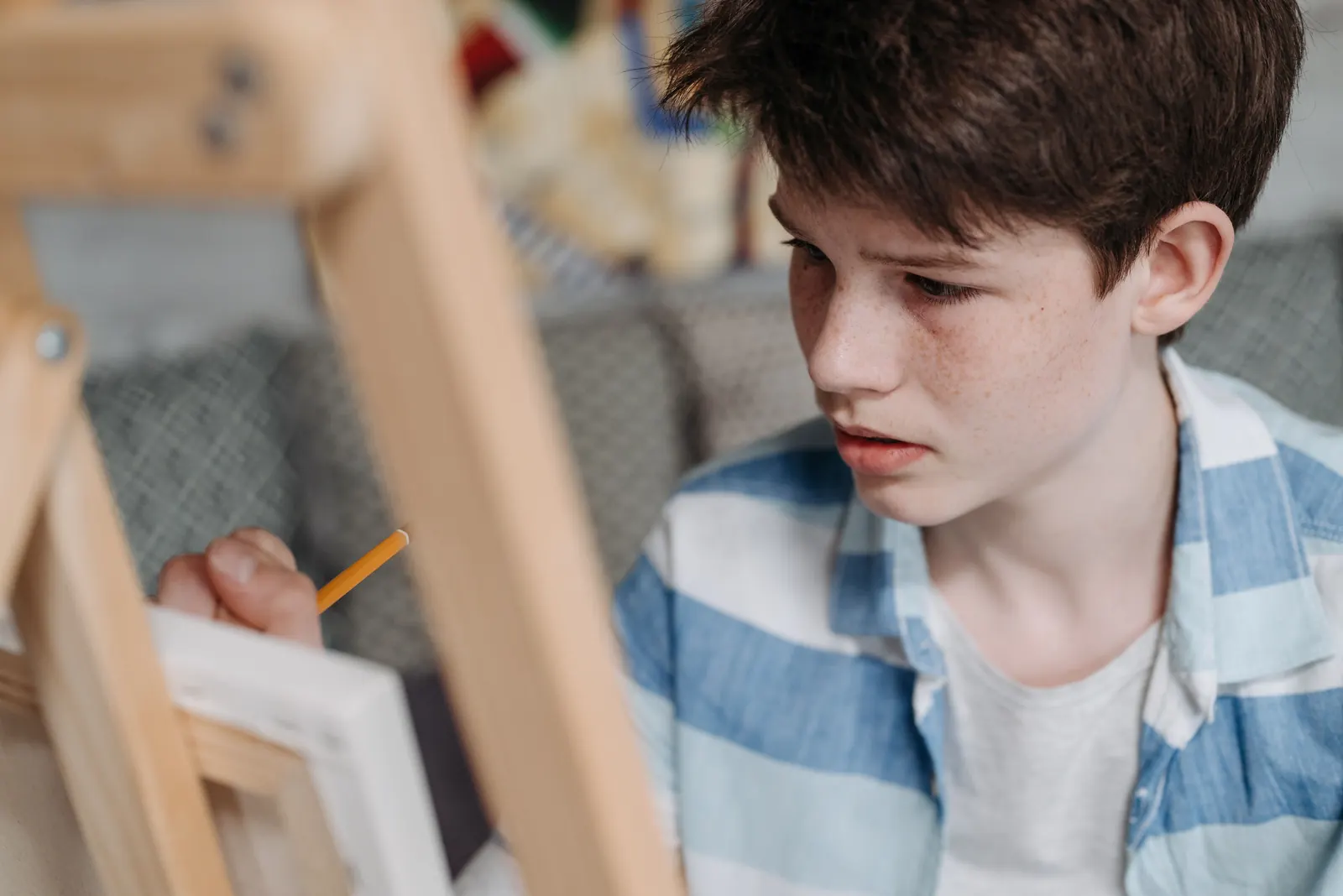 A boy in a blue striped shirt sketches with a pencil at a wooden easel, concentrating