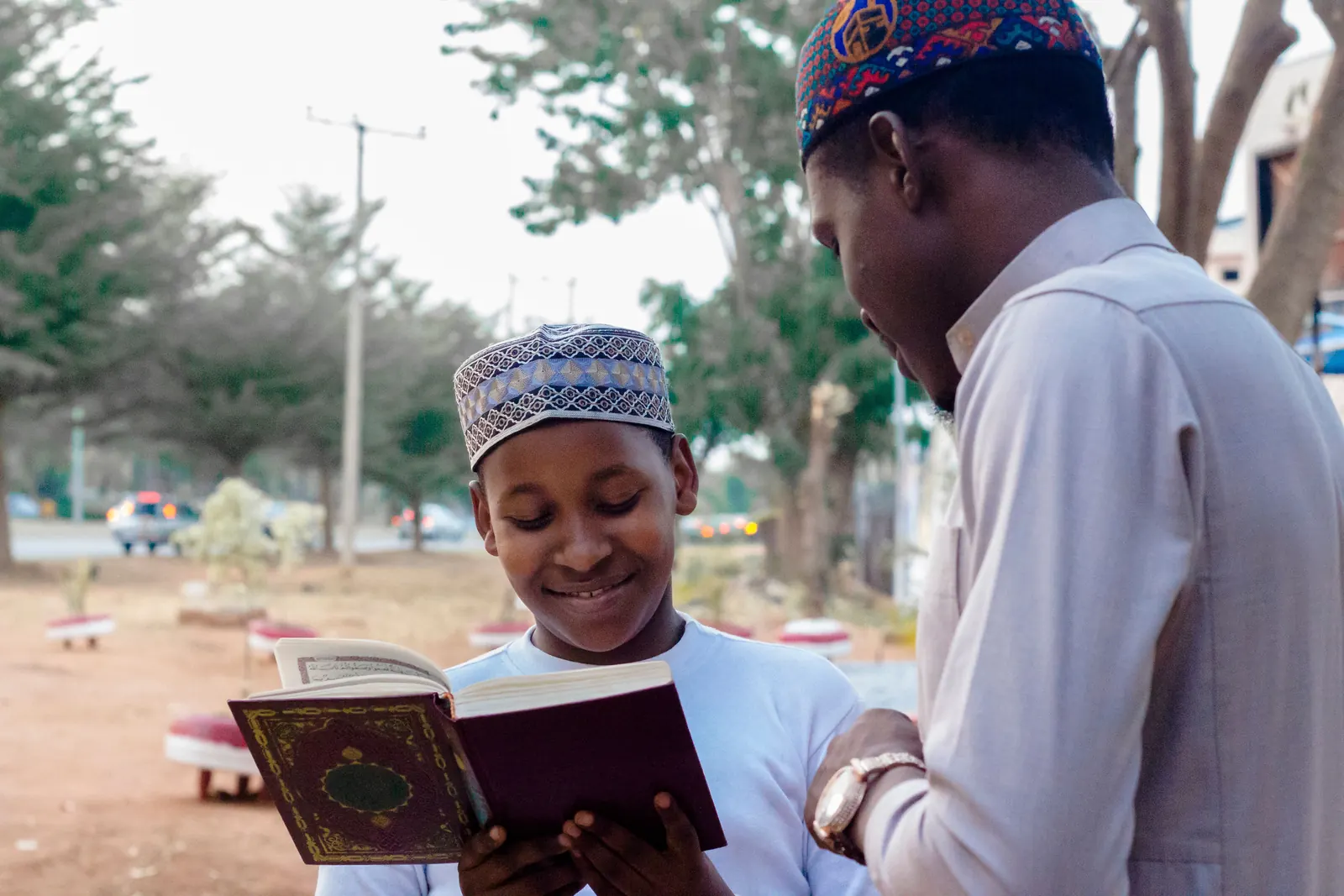 A boy in an embroidered kufi smiles over an open Qur'an while a man in a kufi beside him guides the reading