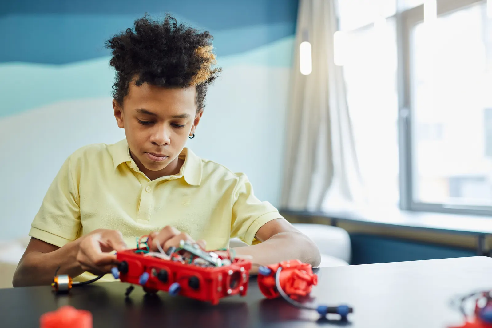 A boy in a yellow polo assembles a red robotics kit with wires and motors at a classroom table