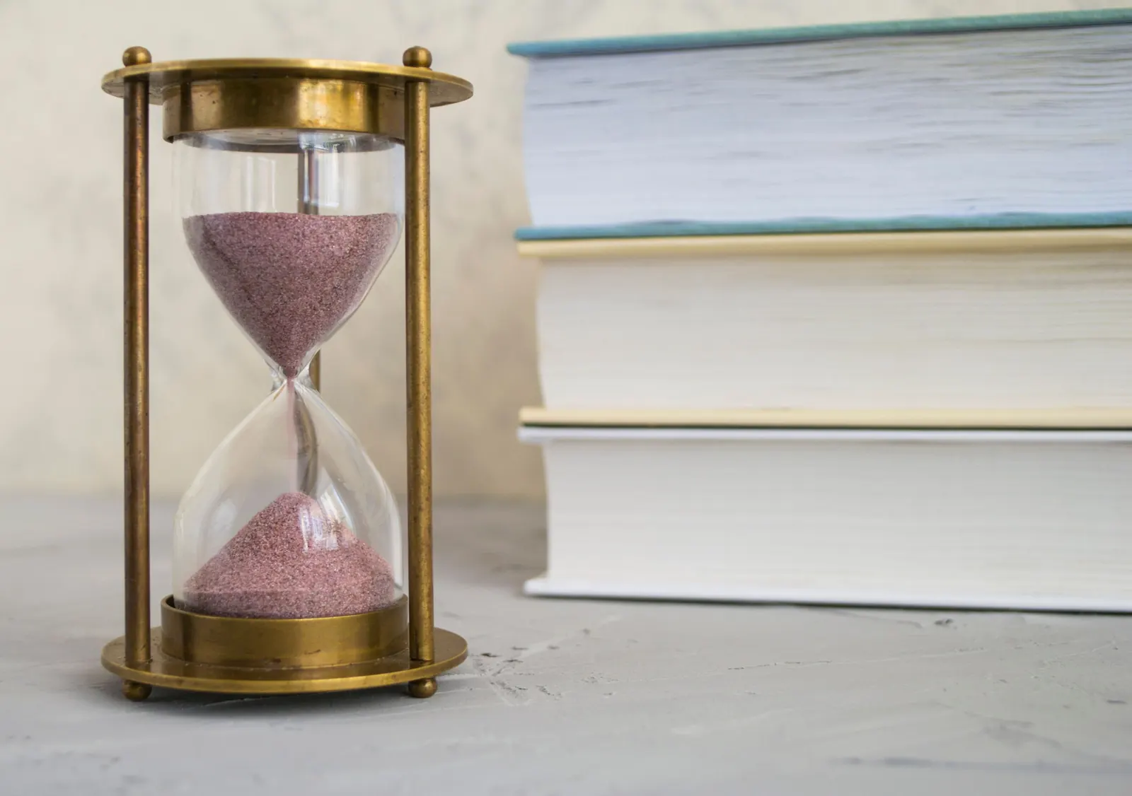 A brass hourglass with pink sand beside a stack of closed hardback books on a grey surface