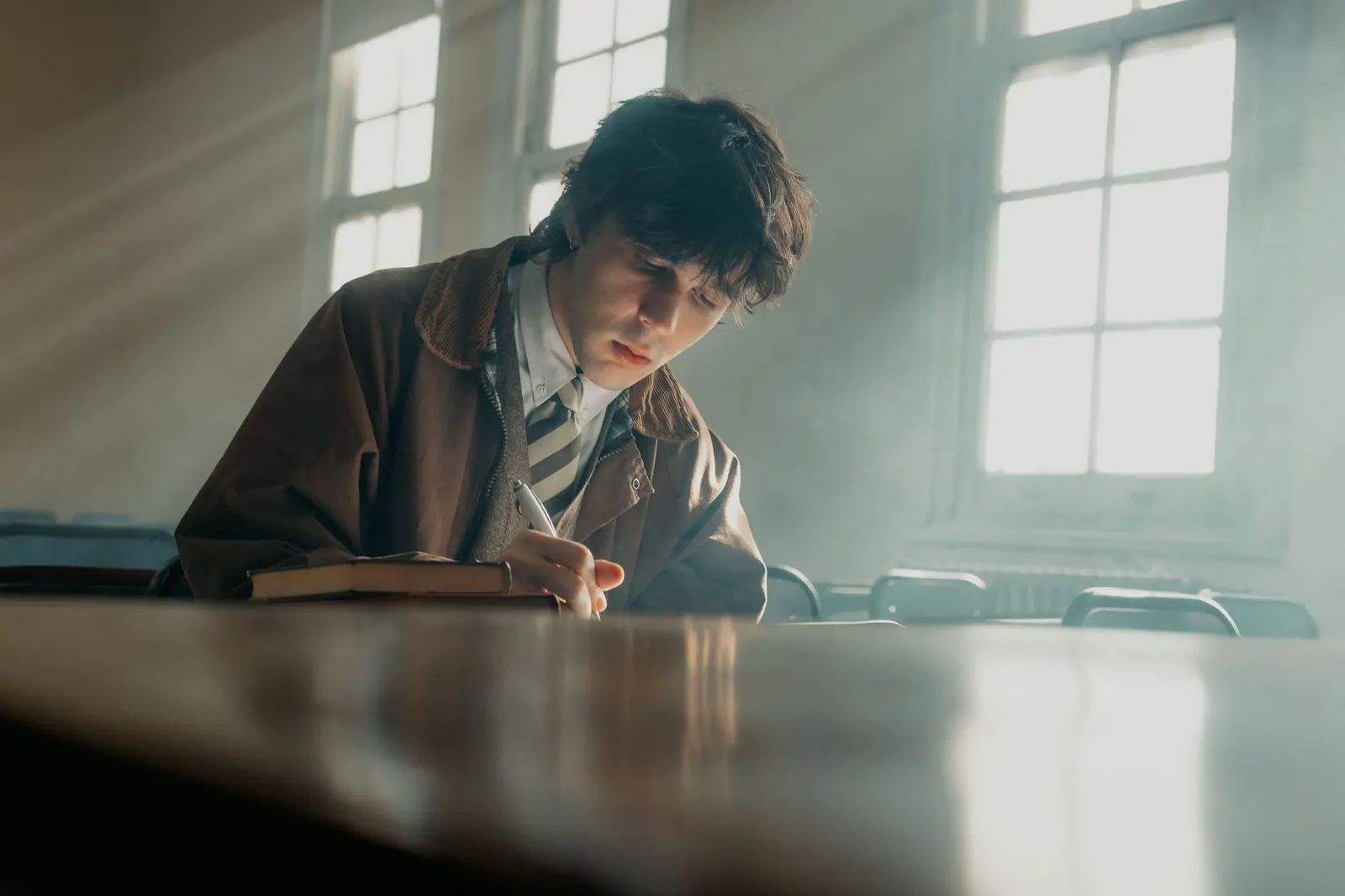 A young man in a jacket and tie writes in a notebook at a desk in a sunlit, empty classroom