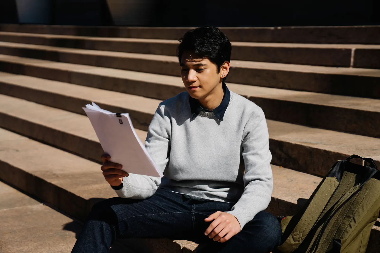 A teenage boy in a grey sweater reads through printed papers on sunny stone steps, backpack beside him
