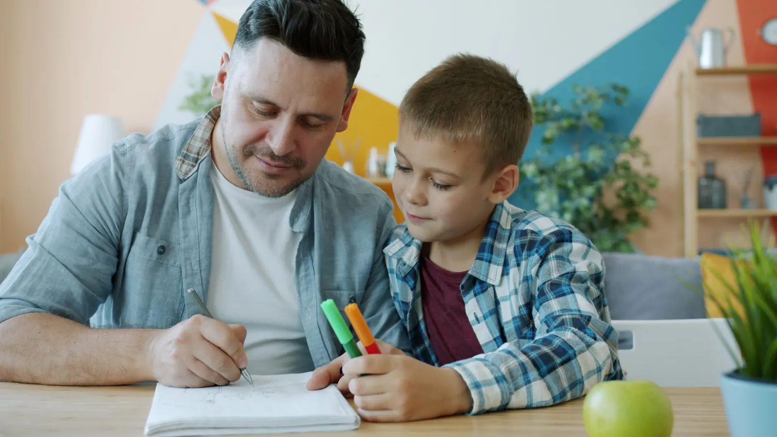 A short-haired man in a denim shirt writes in a notebook beside a boy in a checked shirt holding felt-tip pens at a wooden desk