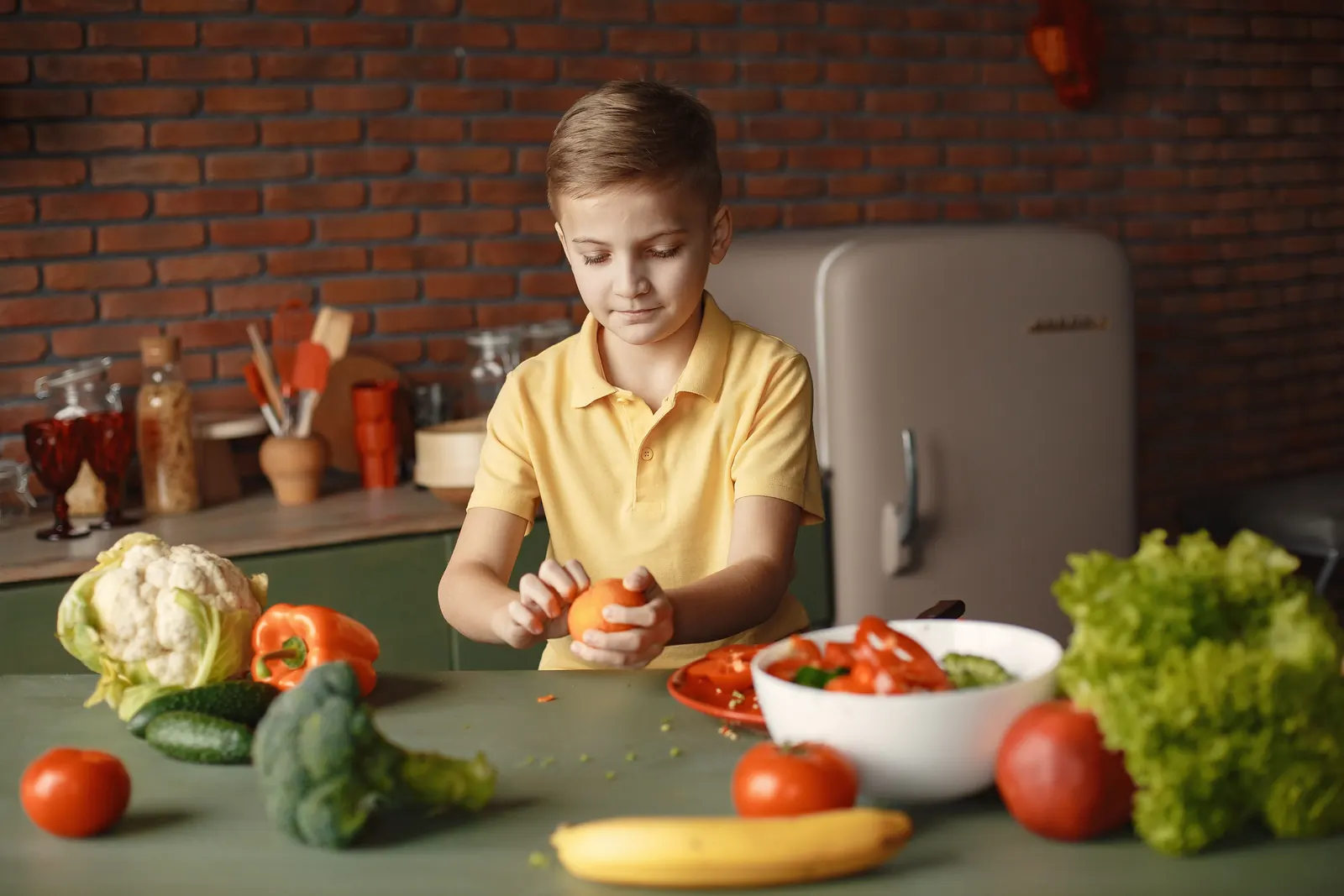 A boy in a yellow polo peels an orange at a kitchen table laid with fresh vegetables and fruit
