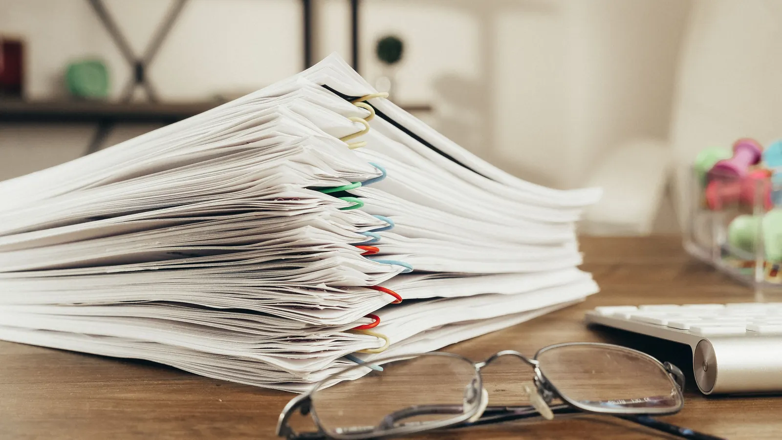 A tall stack of clipped papers and reading glasses on a wooden desk