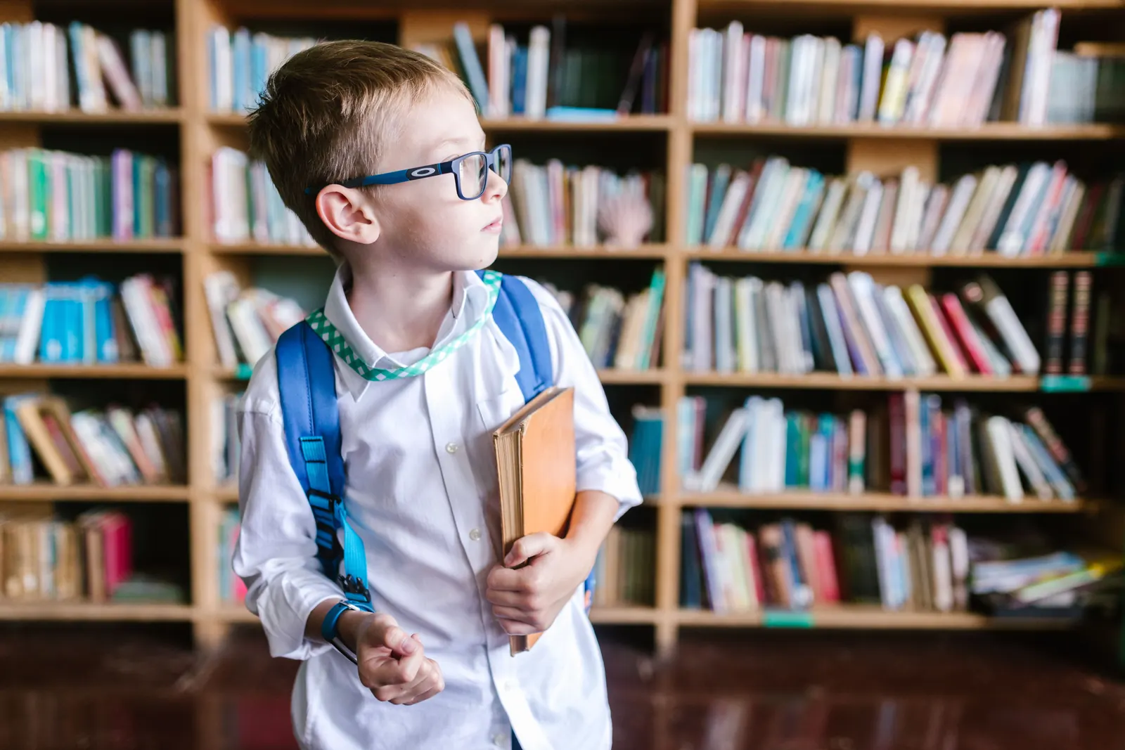 A boy in glasses with a blue backpack holds a book in a school library