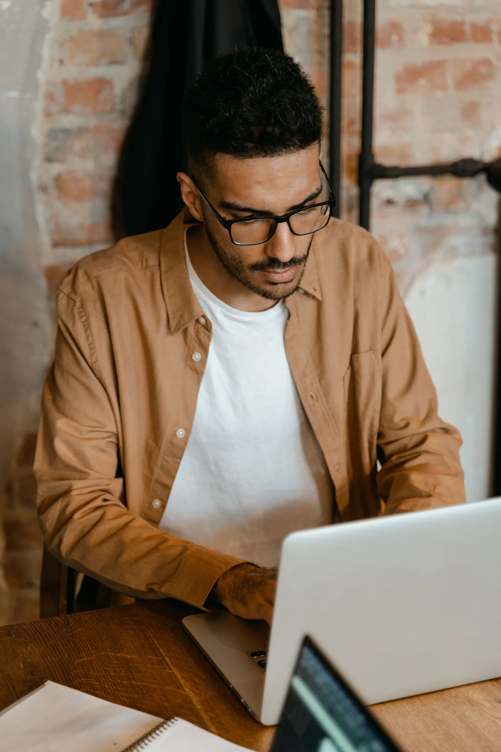 A man in glasses and a tan shirt types on a laptop at a wooden table against a brick wall