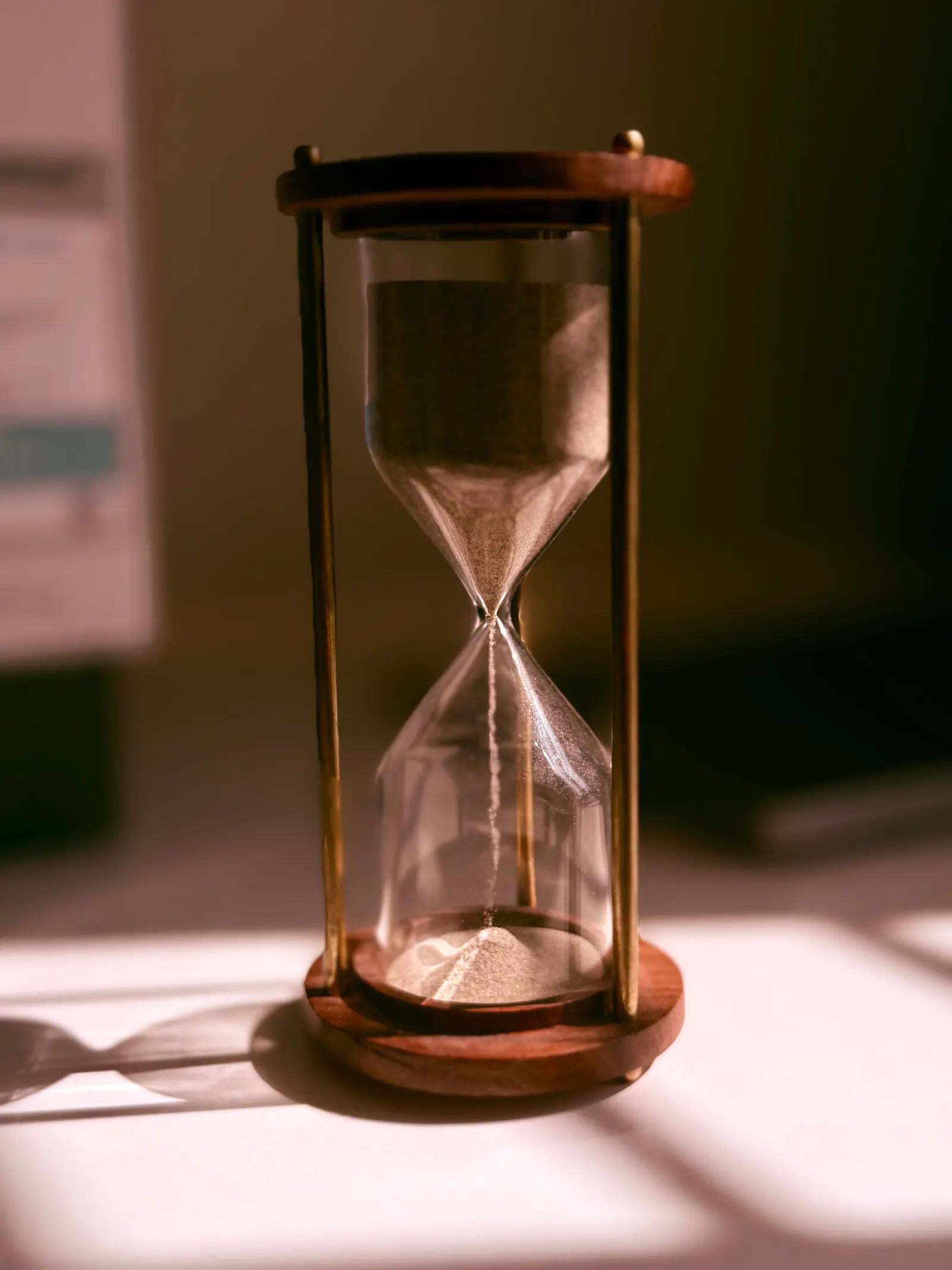 A brass and wood hourglass stands on a table in warm light, sand running through