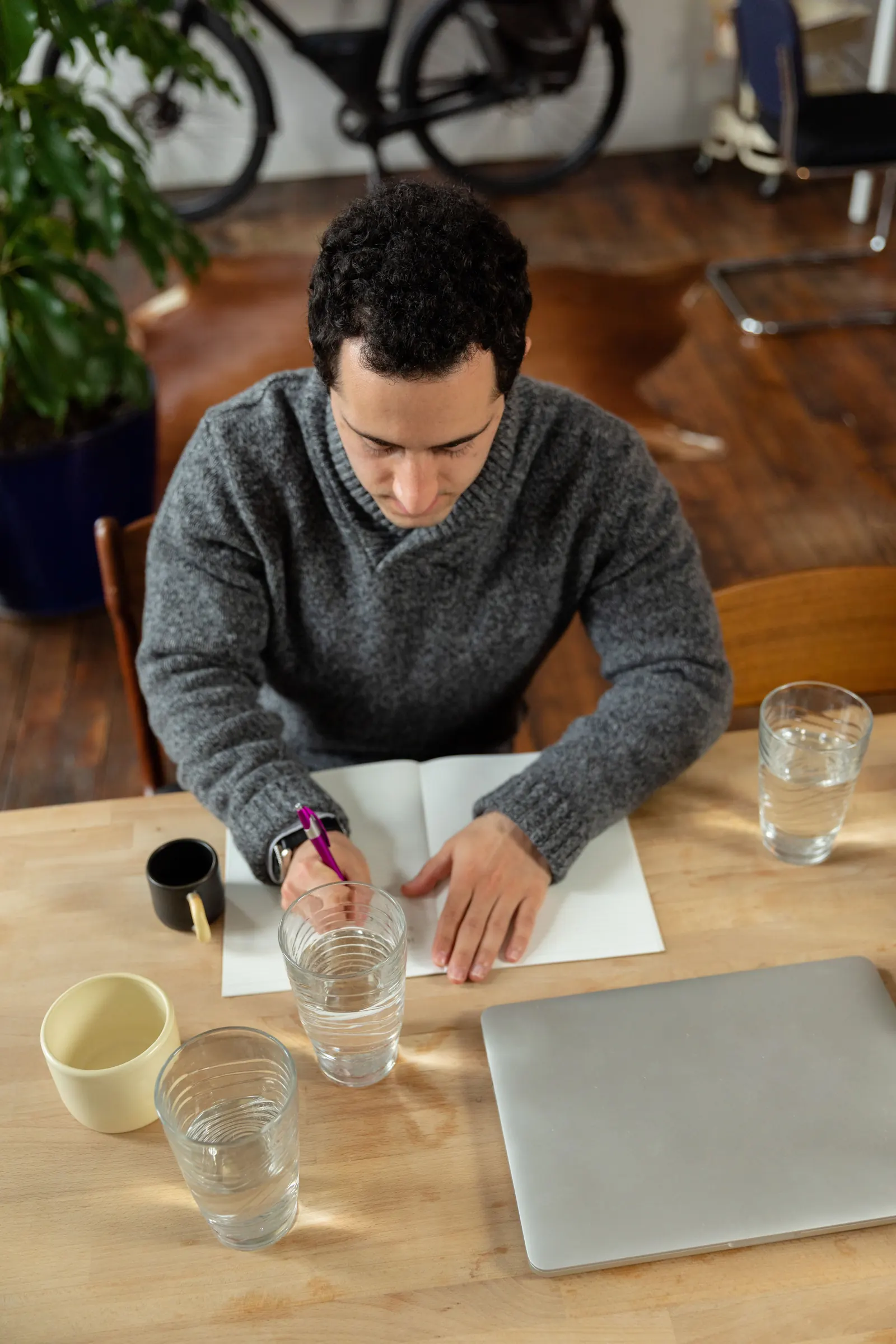 From above, a man in a grey sweater writes in a blank notebook at a wooden table with glasses of water