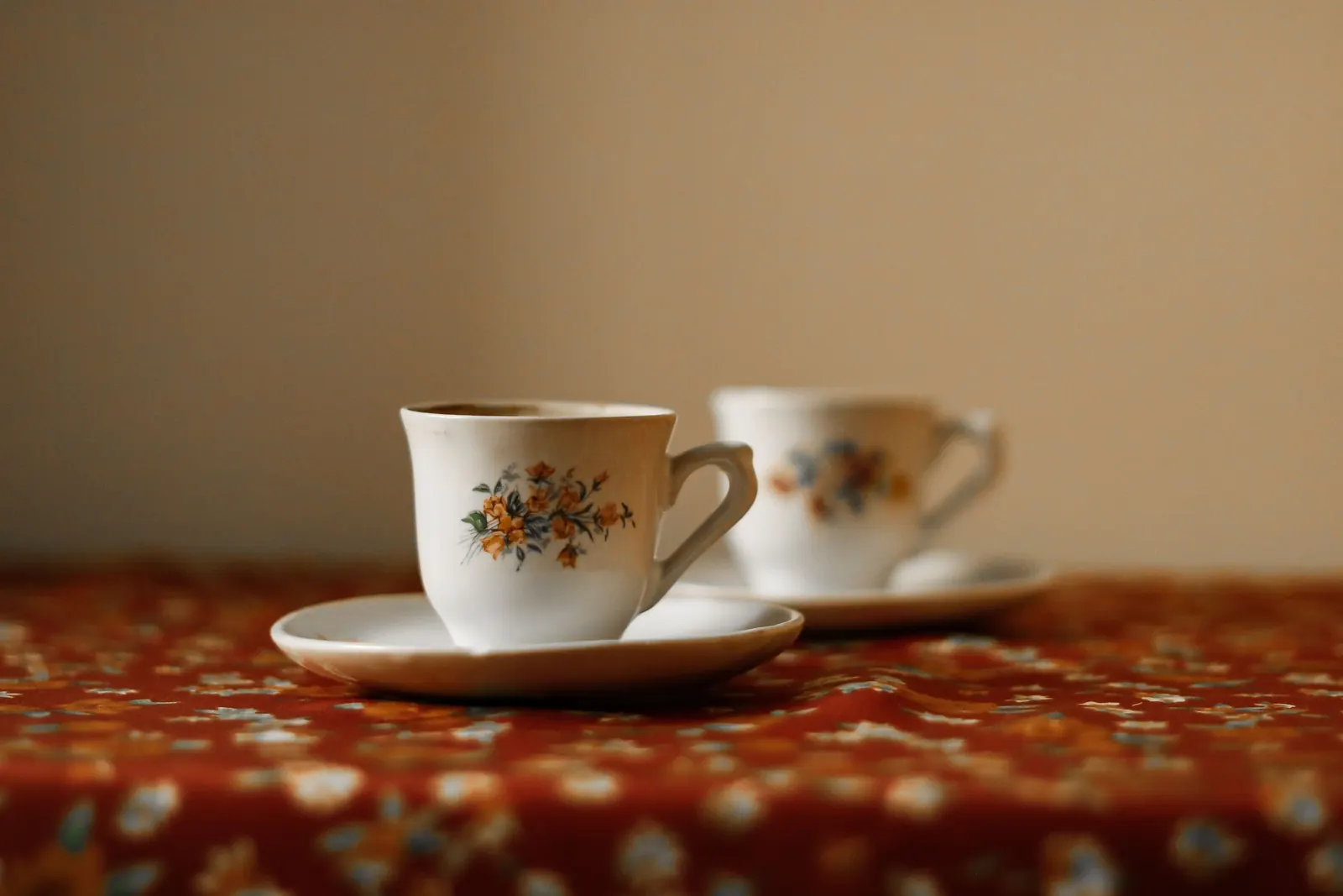 Two white porcelain cups with painted flowers set on saucers on a red floral tablecloth against a warm wall