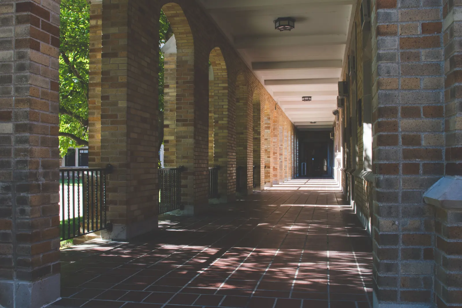 A shaded brick colonnade with arched openings and a tiled walkway leading to a door in dappled sunlight