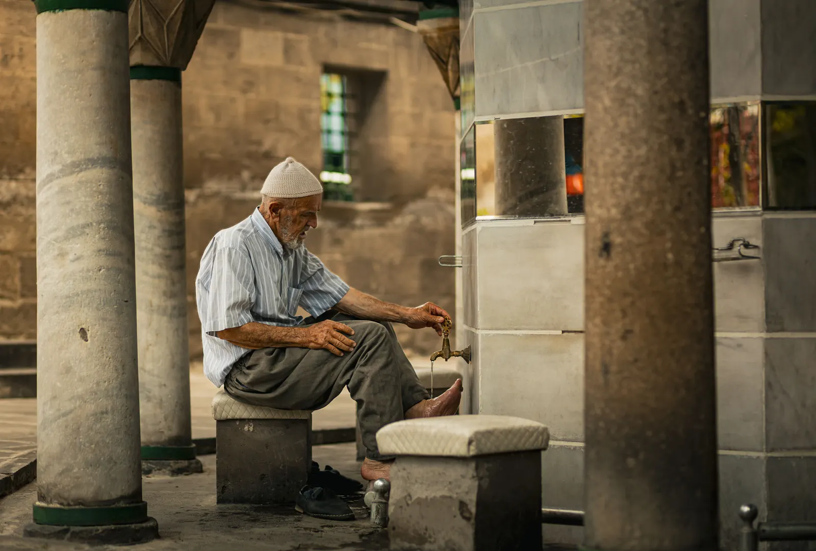 An elderly man in a white cap washes his feet for wudu at a stone fountain between columns