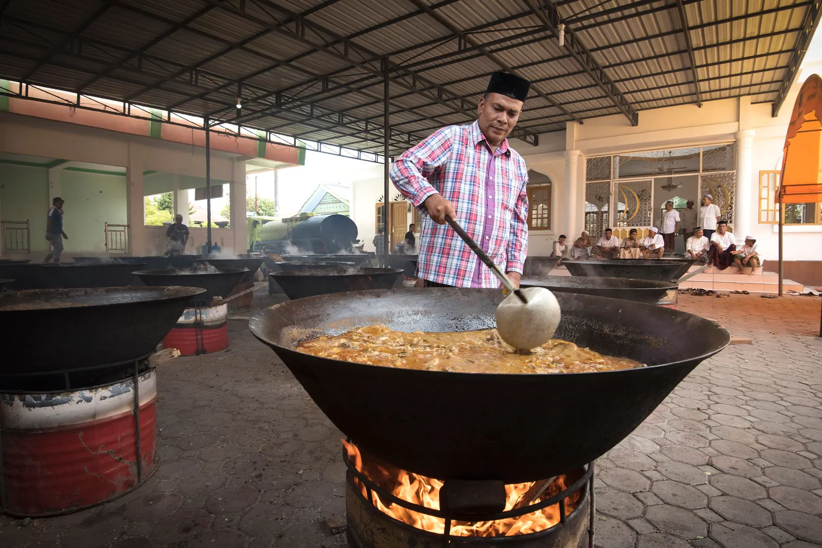 A man in a check shirt stirs a huge pan over a fire while men in caps sit and talk outside a mosque hall
