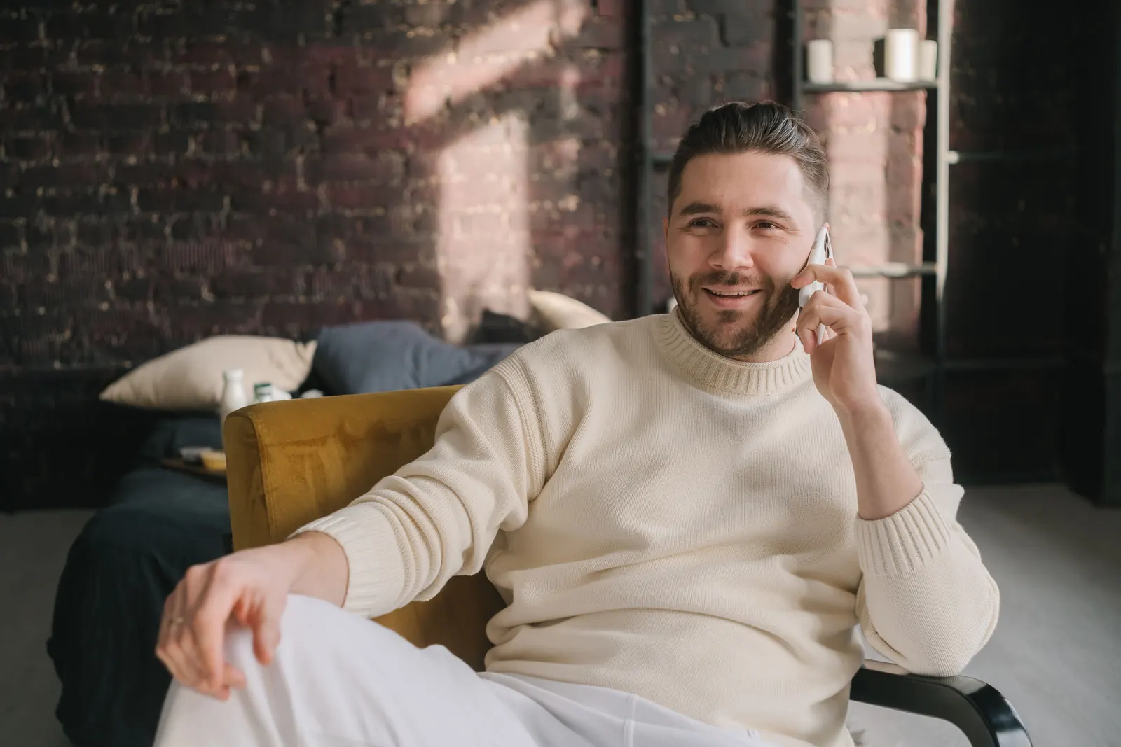 A smiling man in a cream jumper sits in a mustard armchair talking on the phone