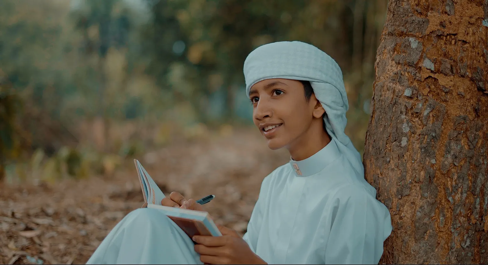 A boy in a pale blue thobe and head wrap writes in a small notebook while sitting against a tree trunk