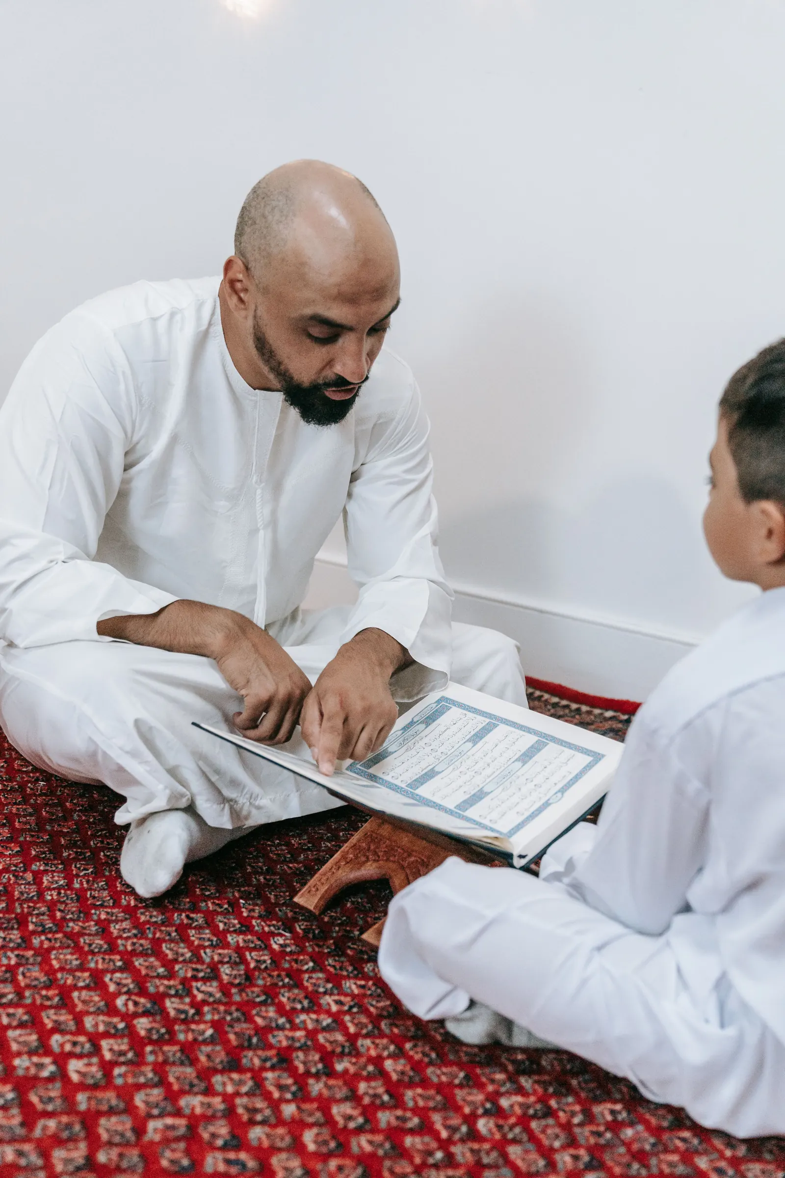 A man in a white thobe points along the lines of a Qur'an on a wooden stand as a boy sits cross-legged beside him