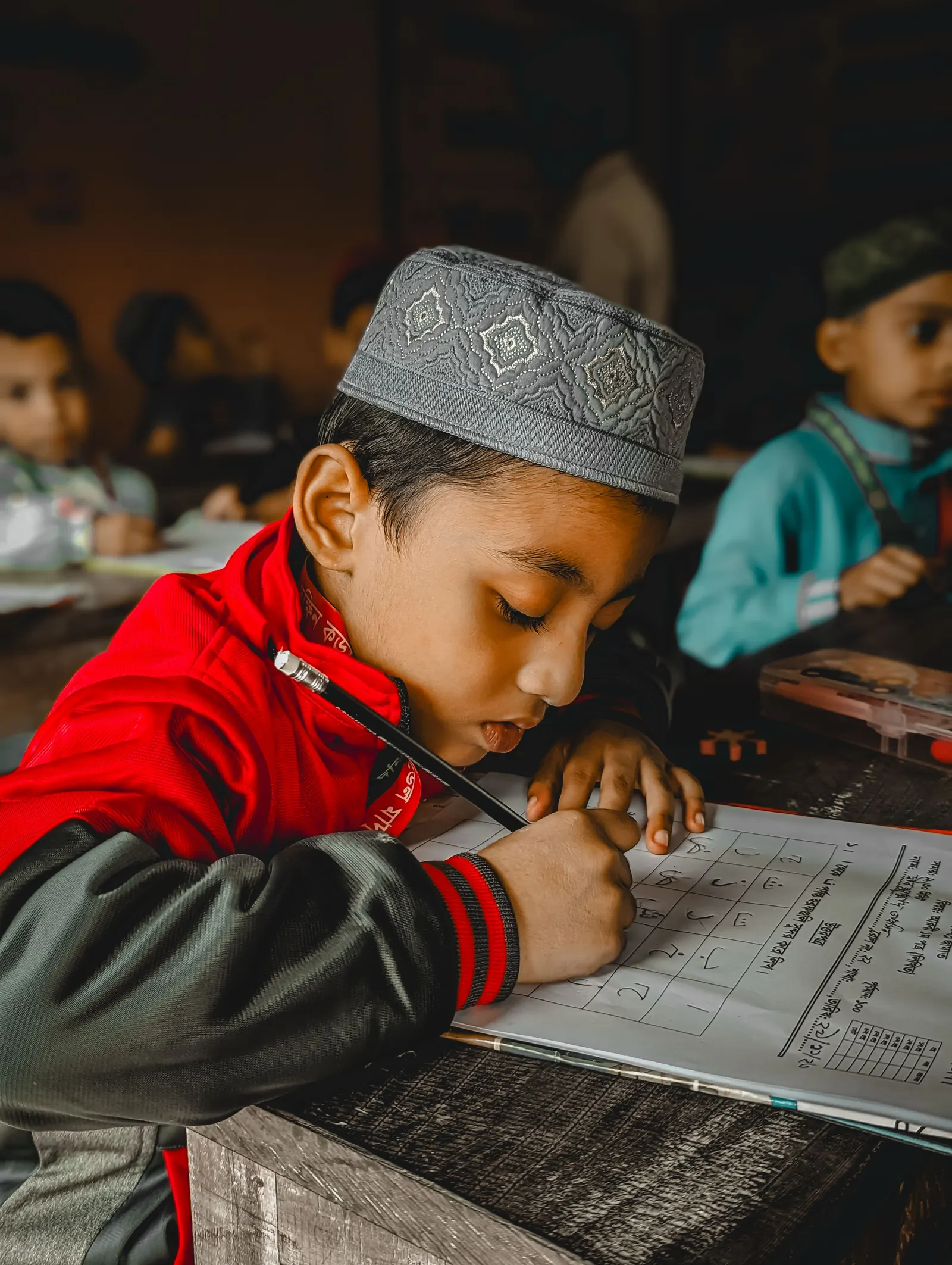A young boy in a patterned kufi bends close over his worksheet, pencil in hand, at a classroom desk
