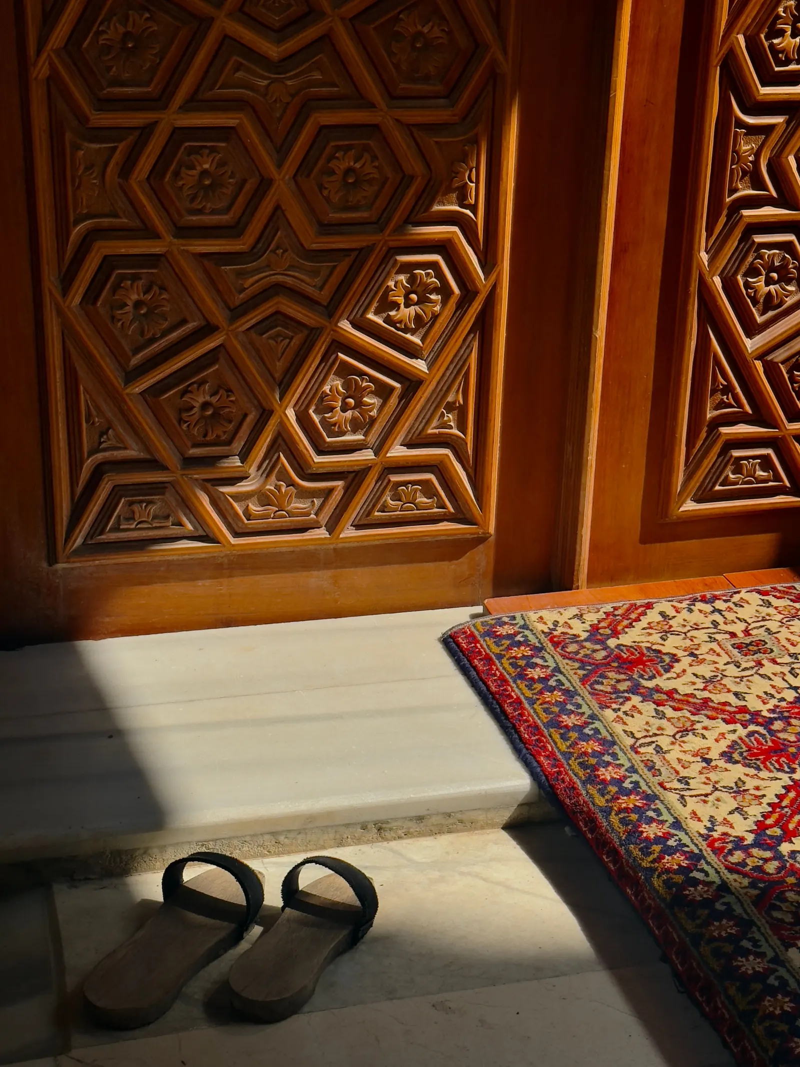A pair of wooden sandals left on the marble threshold beside a patterned rug at a carved mosque door