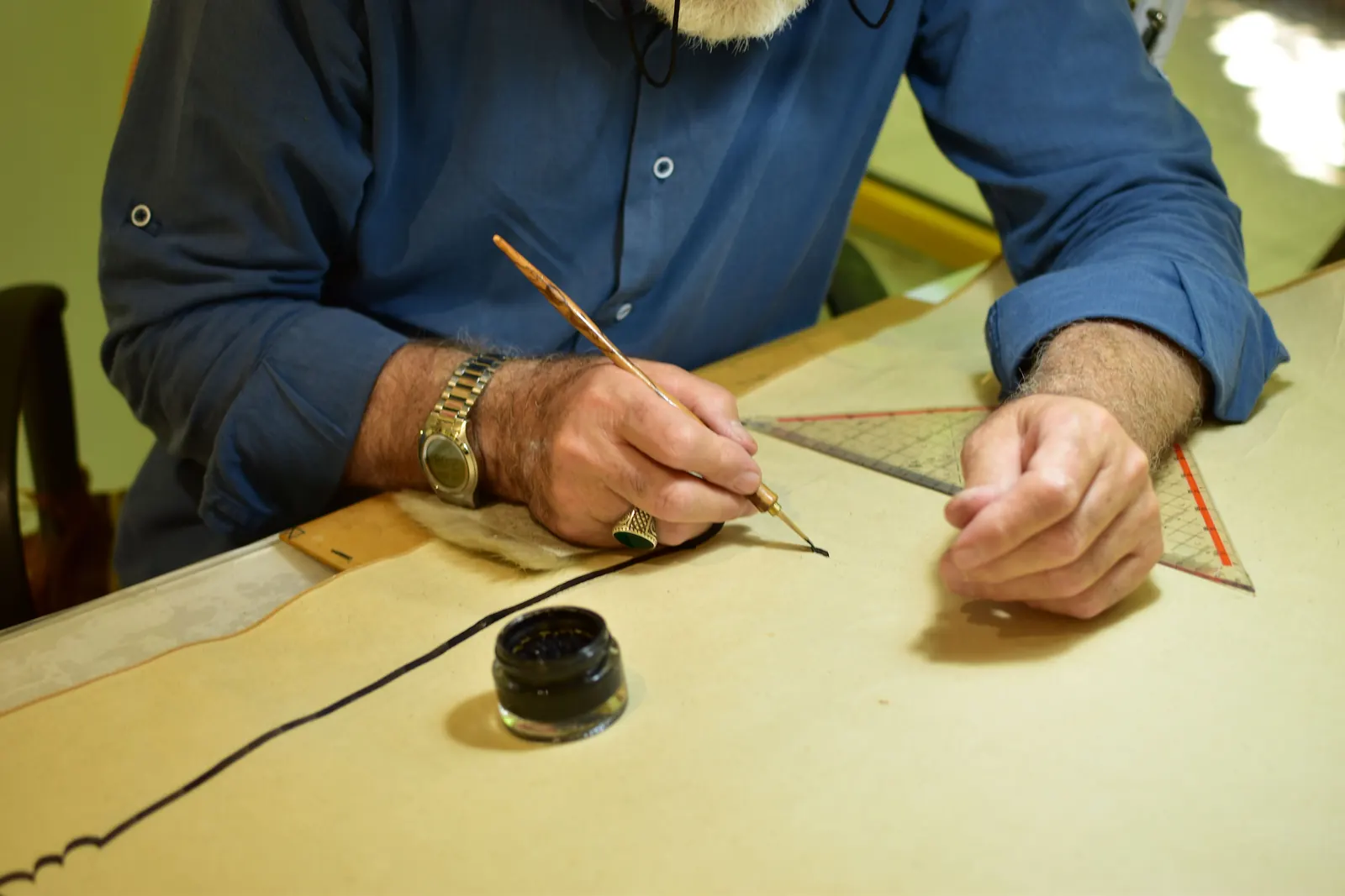 A bearded calligrapher in a blue shirt draws a line with a wooden reed pen beside a small pot of ink