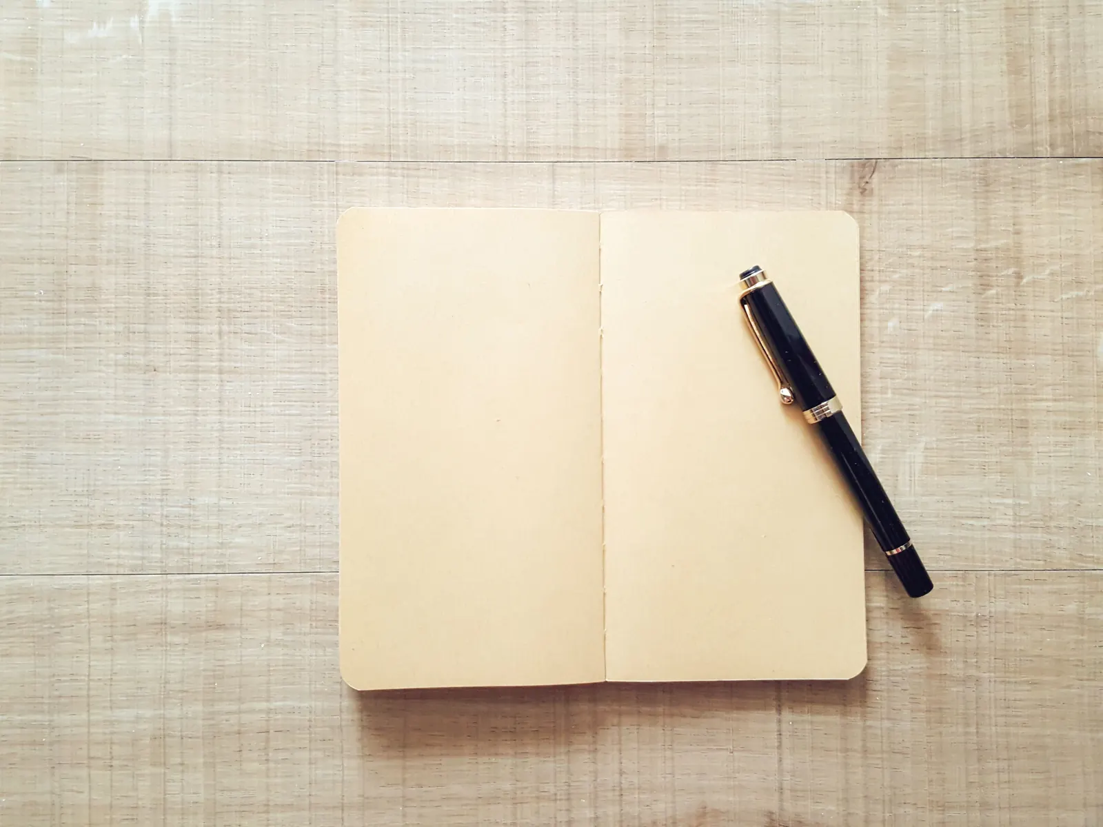 An open blank kraft notebook with a black fountain pen resting on it, on a pale wooden desk