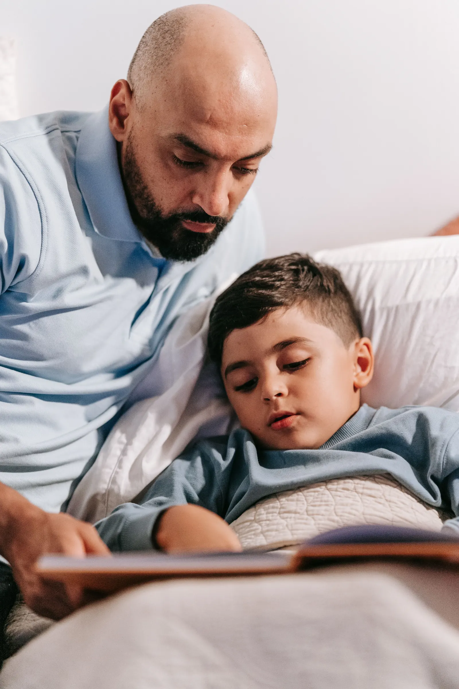 A father sits beside his son tucked into bed and reads to him while the boy rests