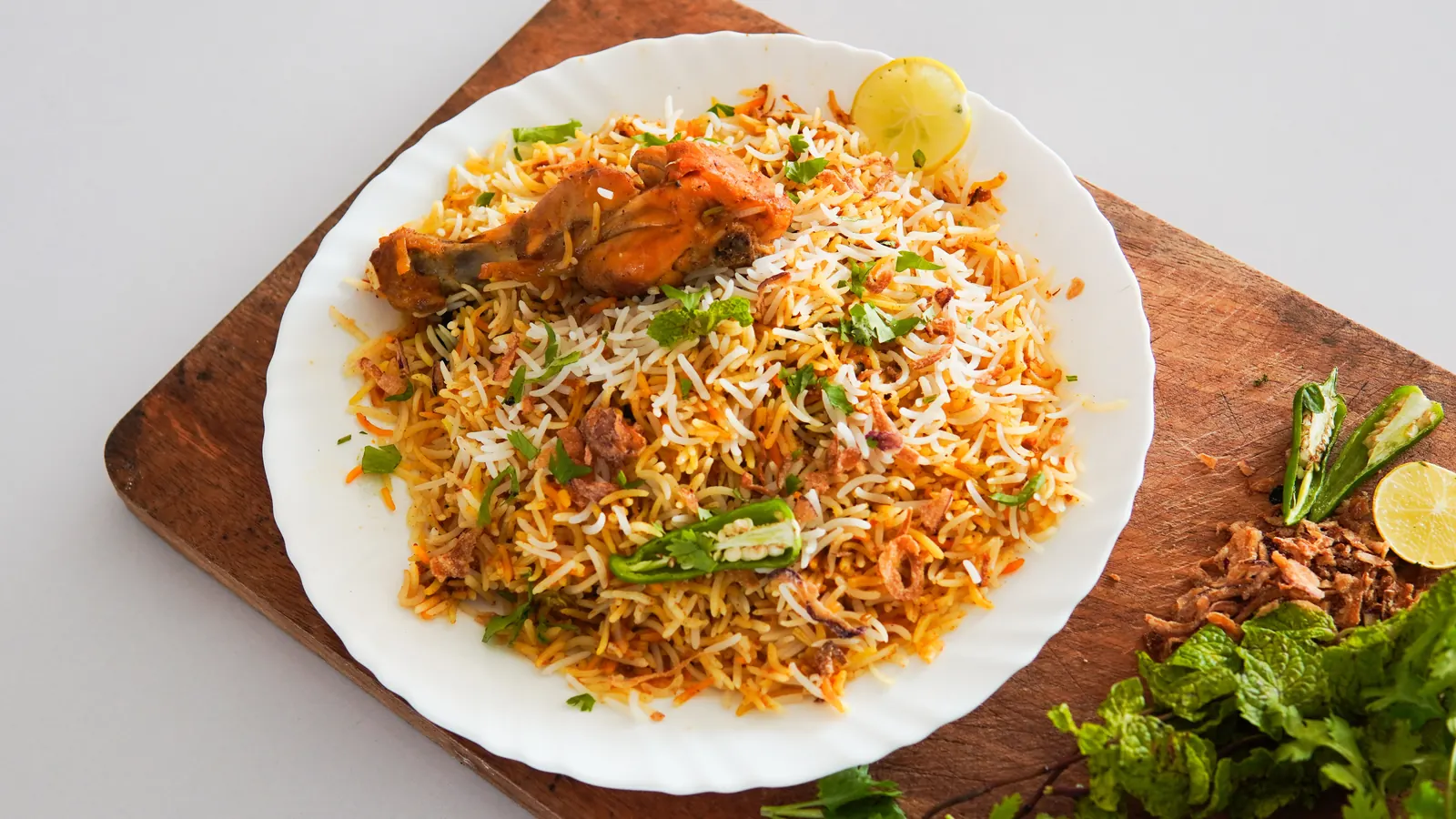 A white plate of chicken biryani with lime, green chilli and mint on a wooden board