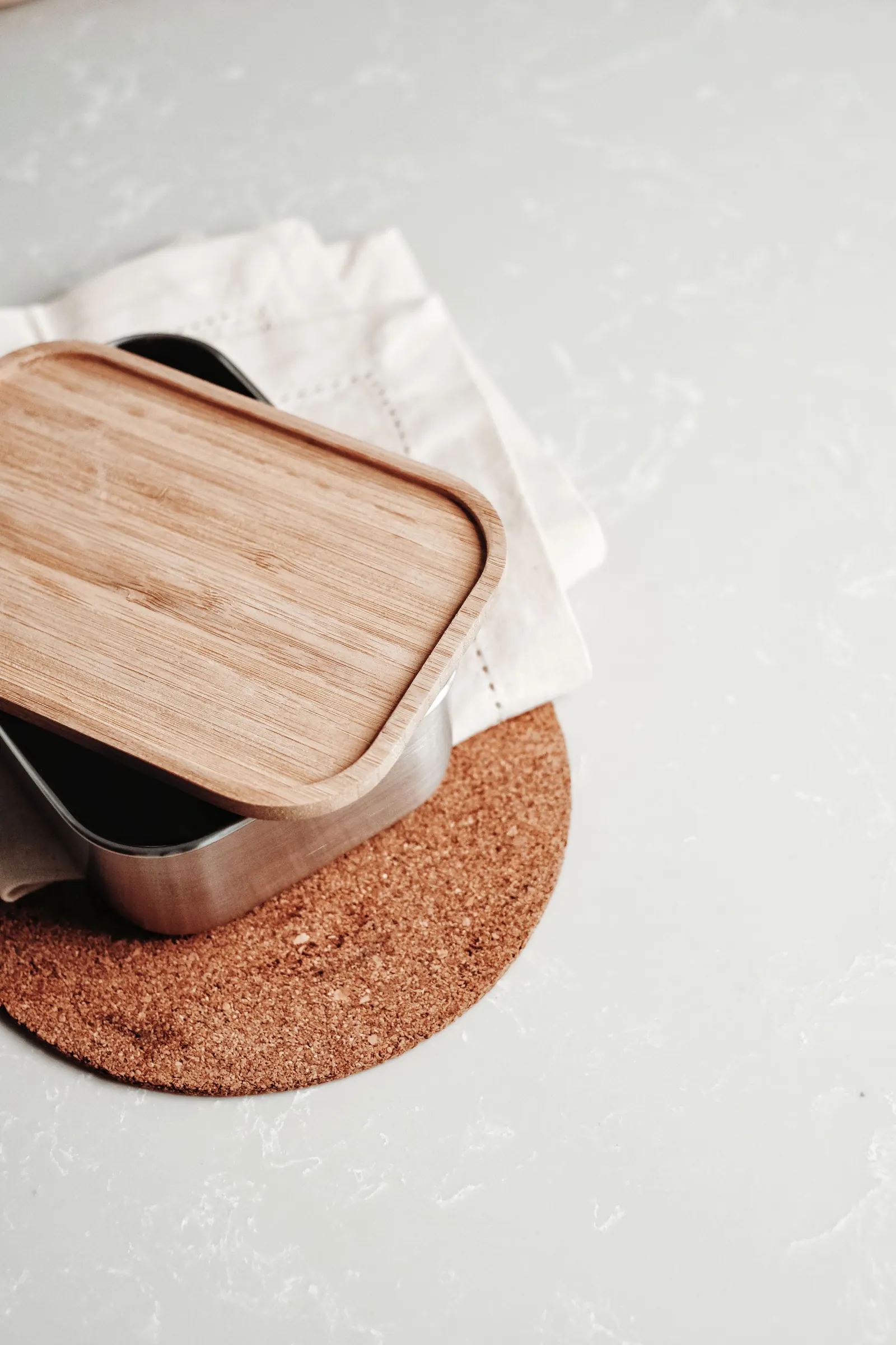 A steel lunch box with a bamboo lid resting on a cork mat and linen napkin