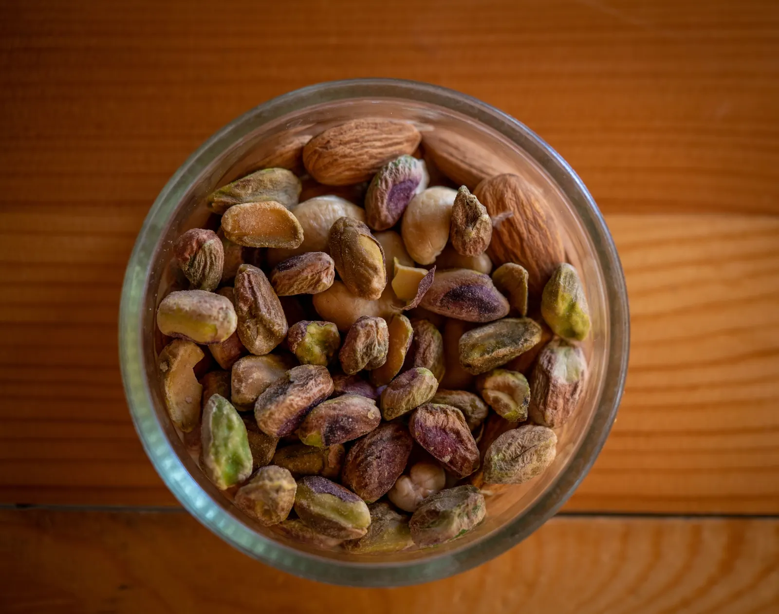 A glass bowl of pistachios and almonds seen from above on an orange wooden table
