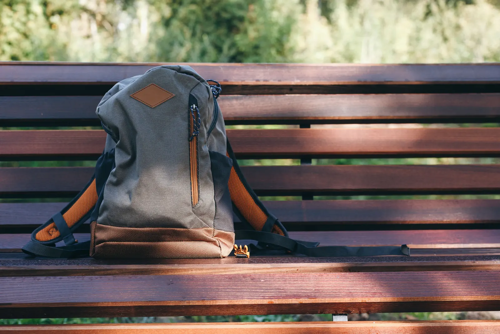 A grey and brown backpack left on a slatted wooden bench in dappled sun