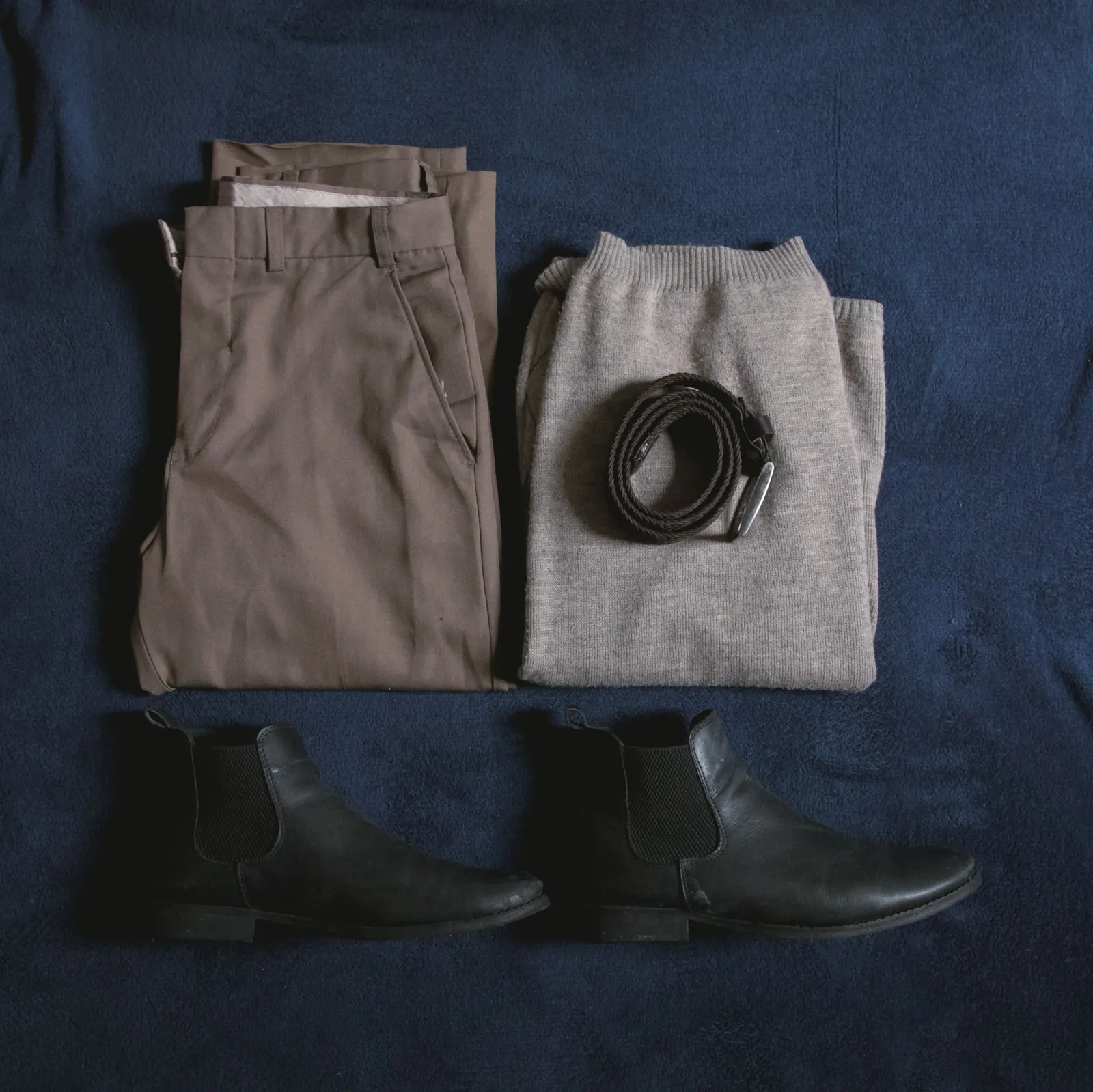 Neatly folded brown trousers, a grey jumper, a belt and black leather boots laid out on navy cloth