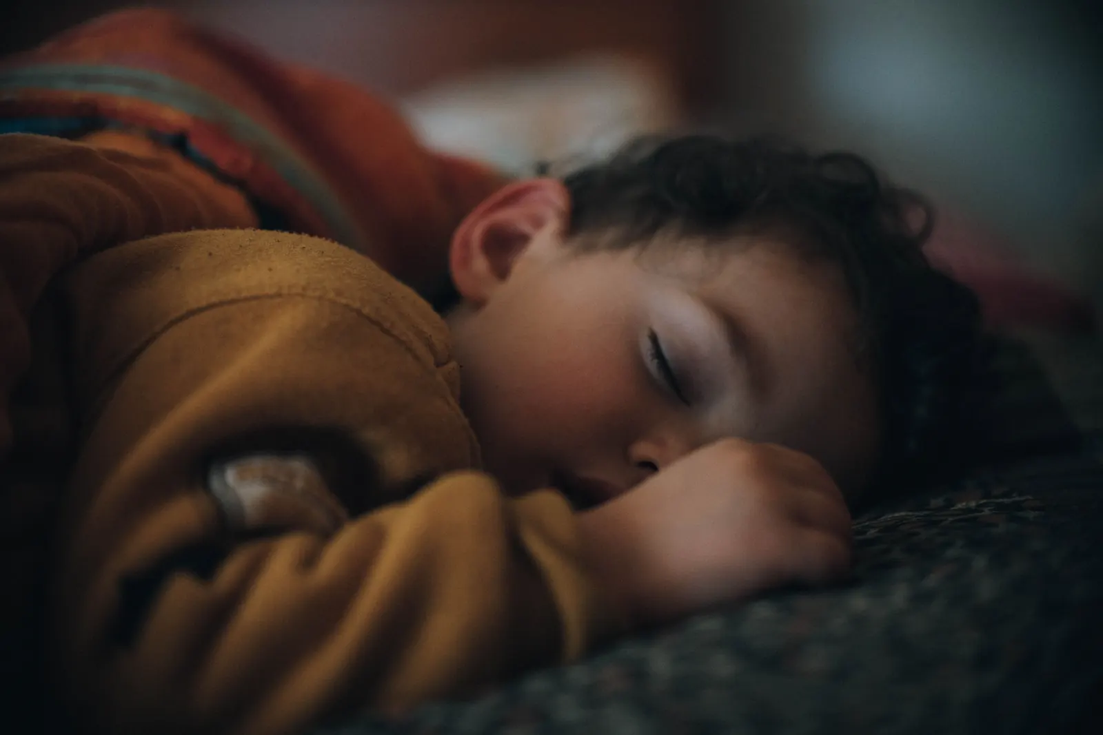 A small boy sleeps peacefully, cheek on his hand, under a mustard blanket