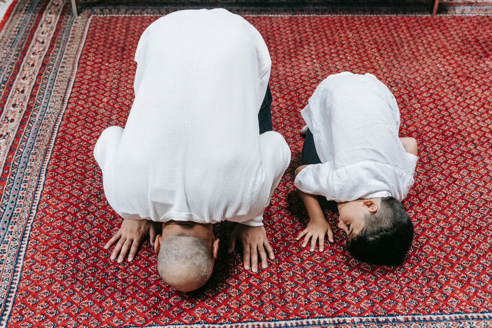 A man and a young boy in white prostrate side by side on a red prayer rug