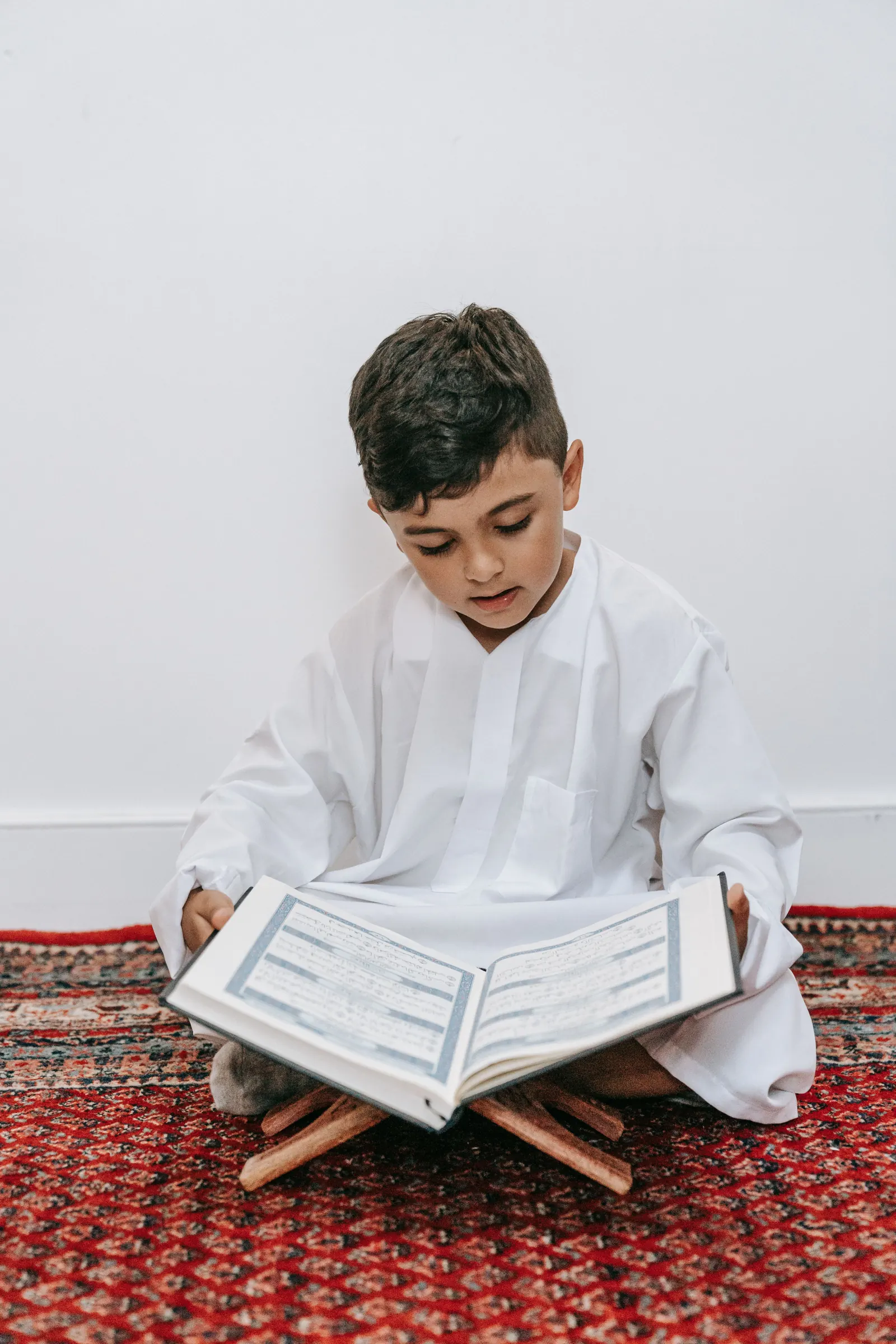 A boy in a white thobe sits cross-legged on a red rug reading aloud from a Qur'an on a wooden stand