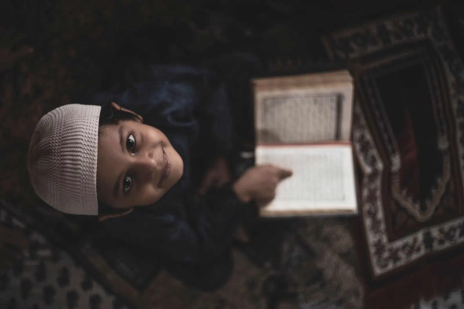 A smiling boy in a white knit cap looks up from an open Qur'an on a patterned prayer mat