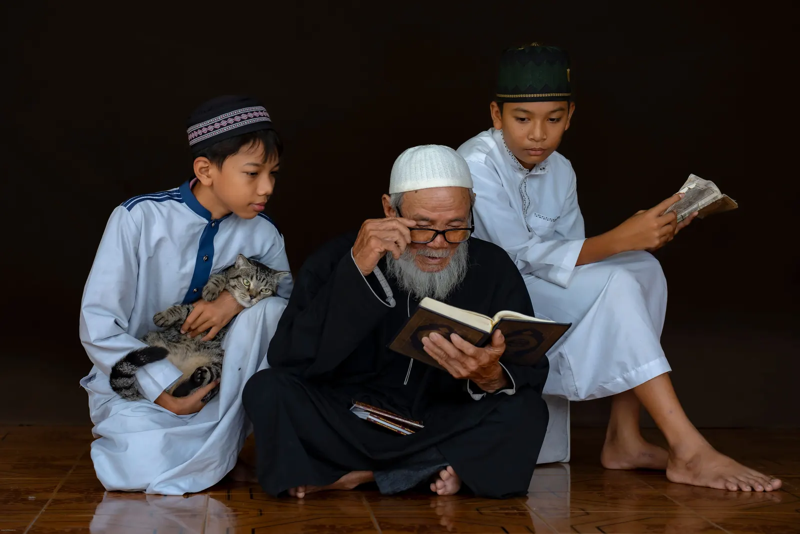 An elderly bearded teacher in a white cap reads from a Qur'an as two boys in white thobes and kufis lean in to follow