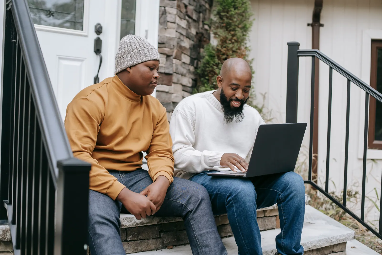 A smiling bearded man types on a laptop as a teenage boy in a knit cap sits beside him on the steps