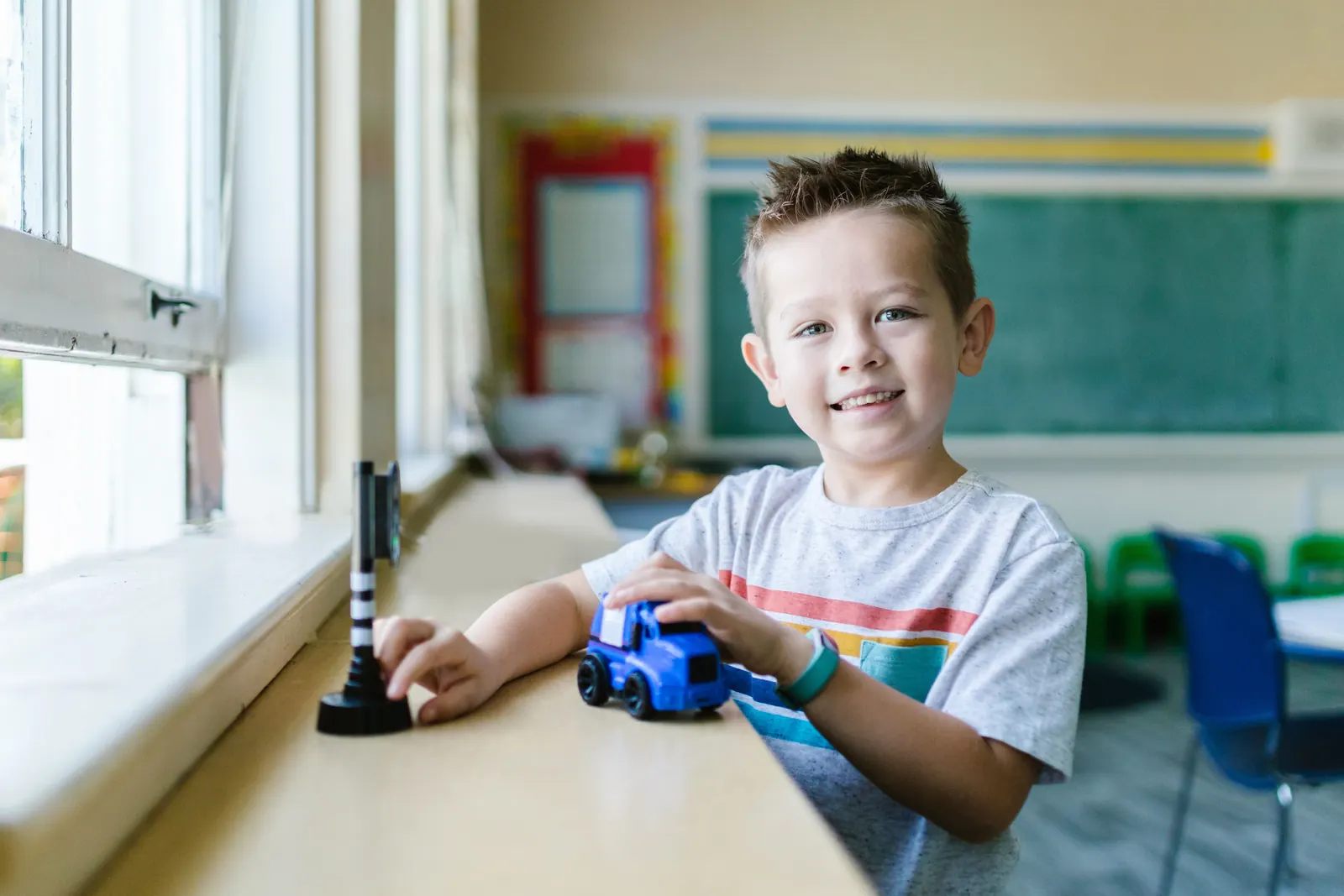 A smiling boy plays with a toy truck on a classroom windowsill