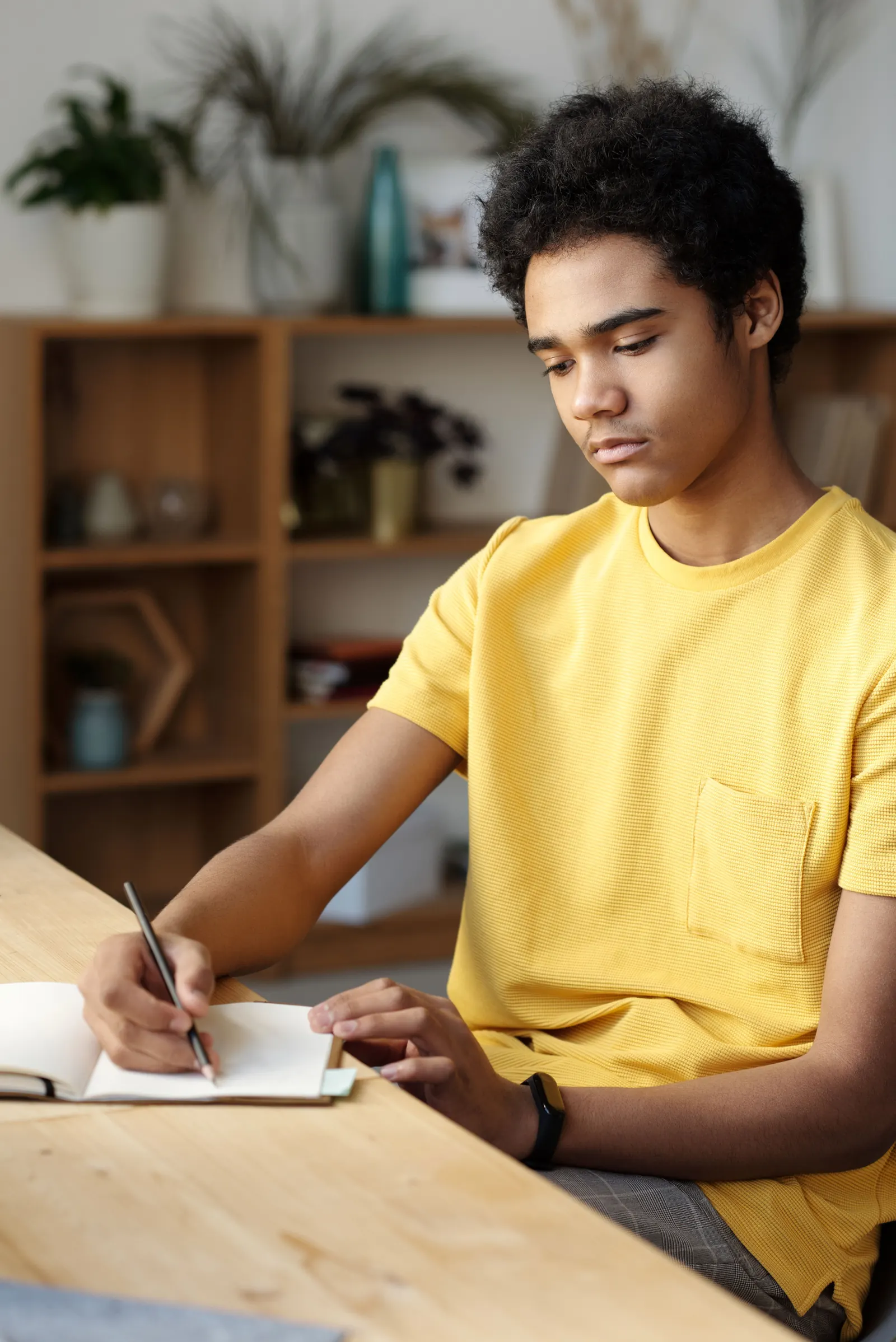 A teenage boy in a yellow shirt writes notes at a desk in a bright room