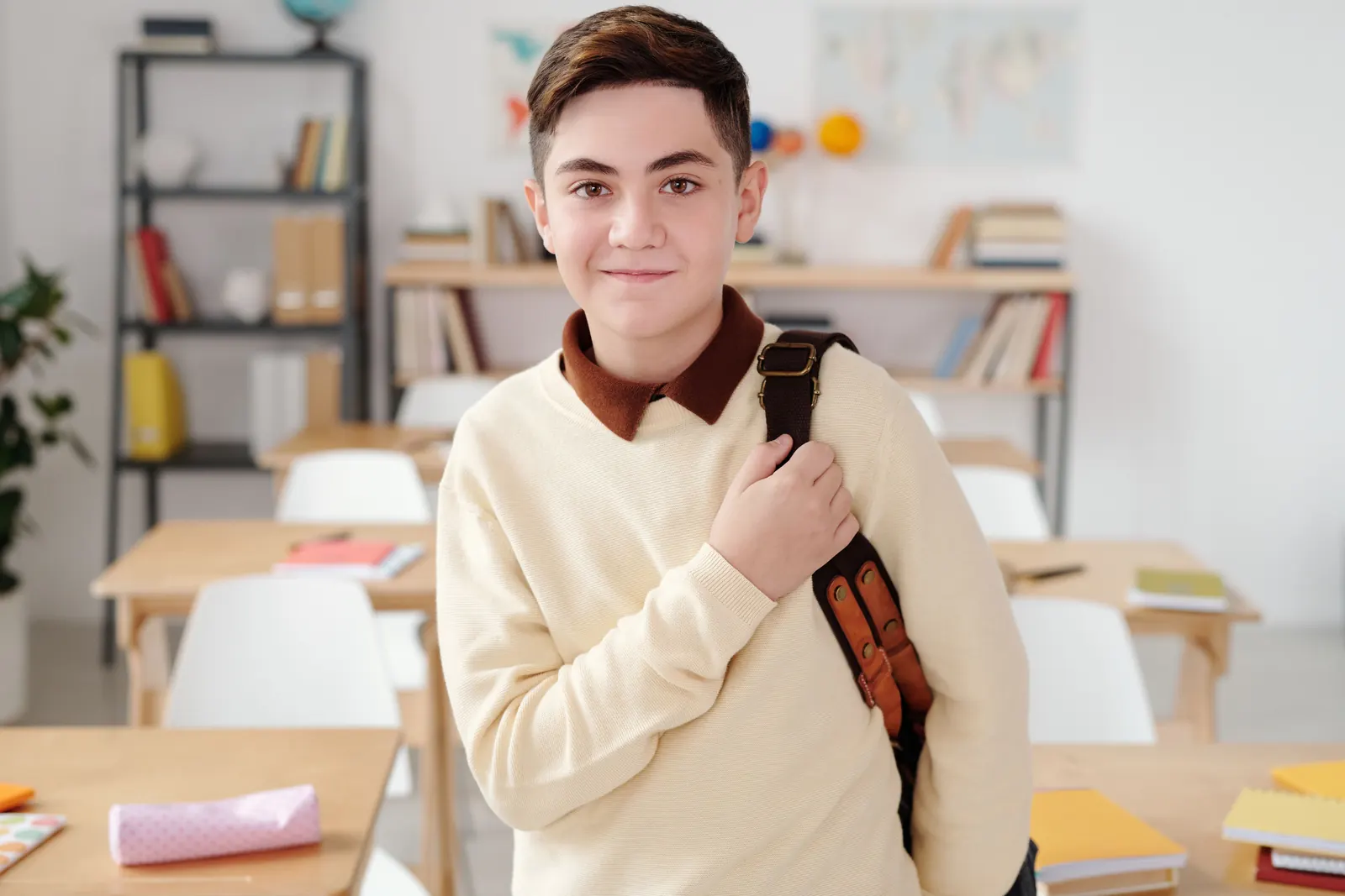 A teenage boy with a satchel over his shoulder smiles in a bright classroom