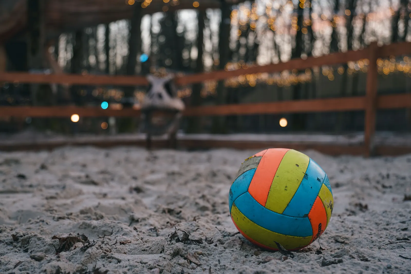A worn volleyball in orange, yellow and blue resting in sand in front of a wooden fence with warm lights