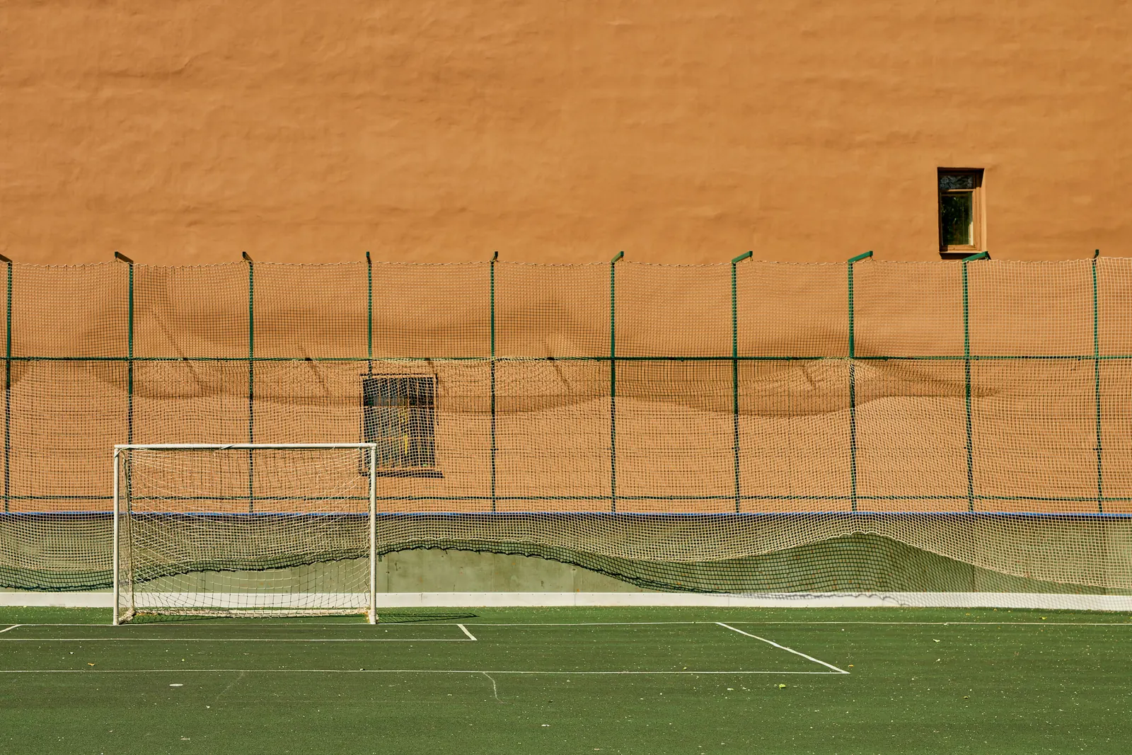An empty small football pitch with a white goal before a high net and a warm terracotta wall