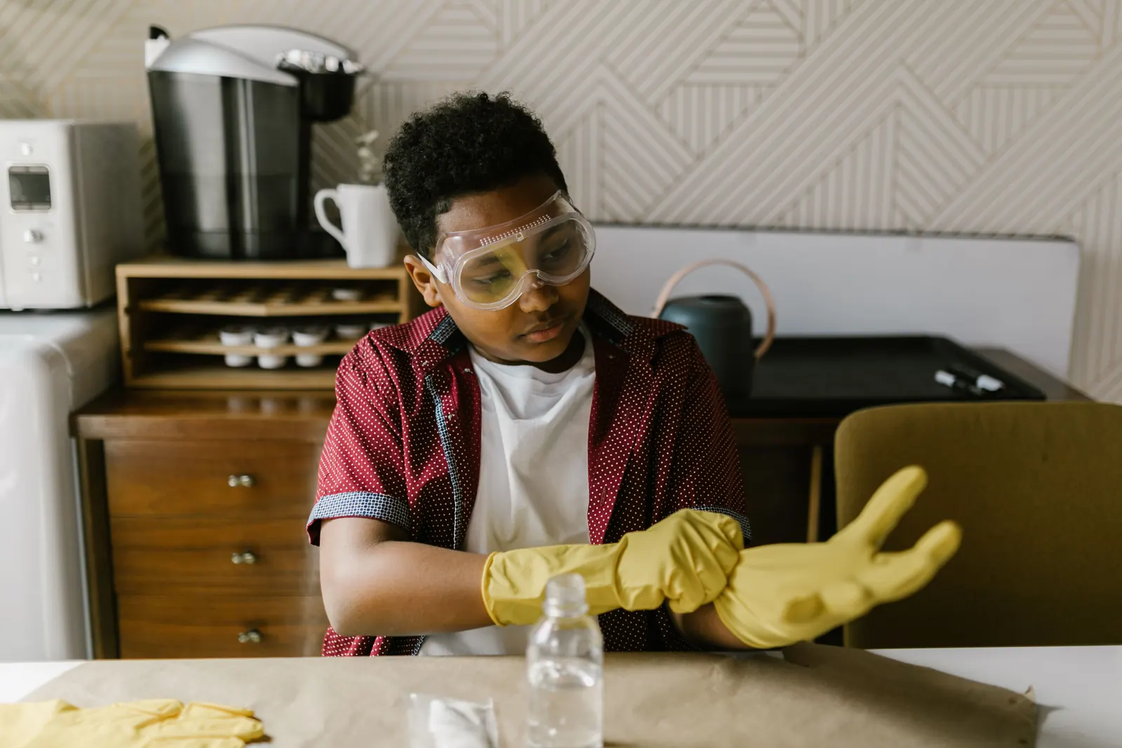 A boy in safety goggles pulls on yellow rubber gloves at a table, ready for a science experiment