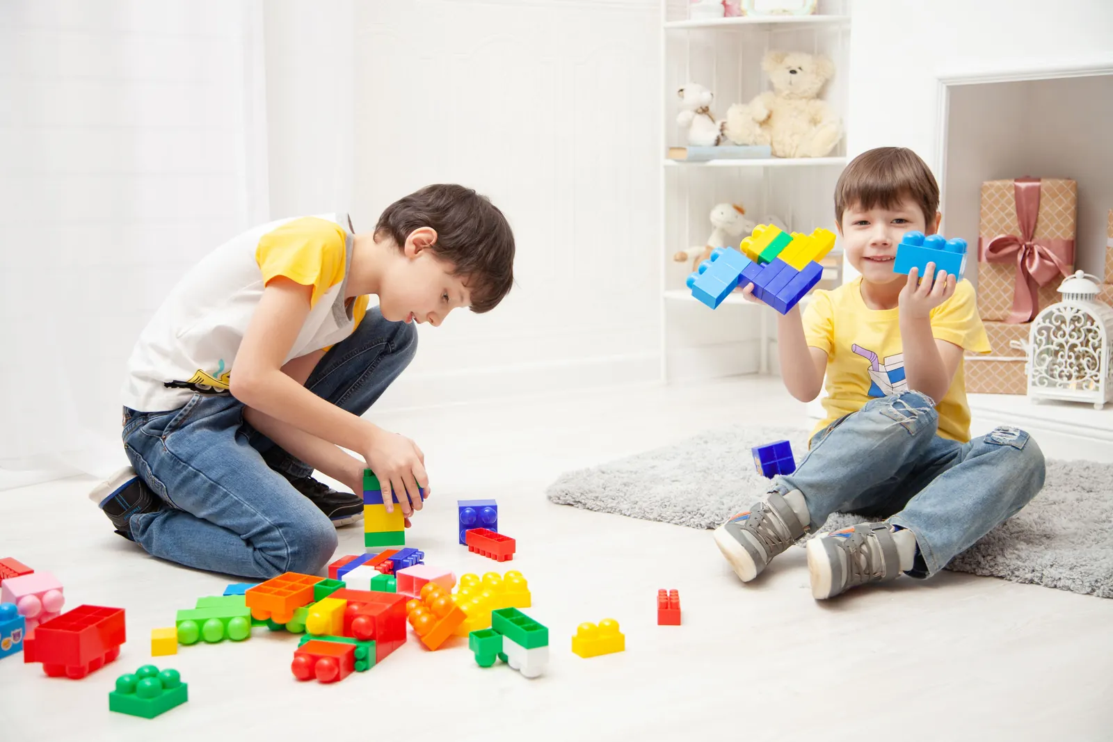 Two young boys build together with bright plastic blocks on the floor of a sunny room