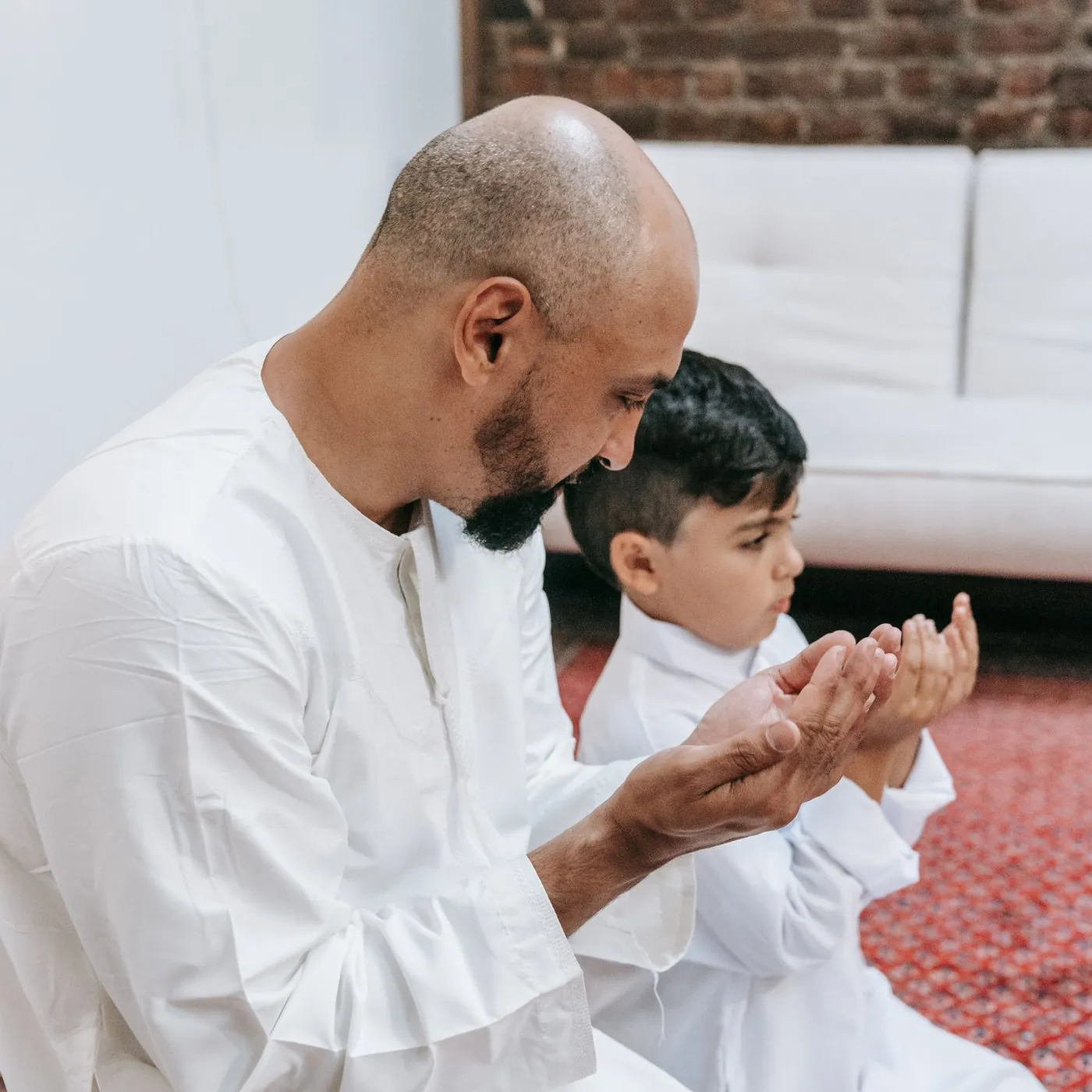 A father and his young son in white thobes kneel side by side on a red prayer rug, hands raised together in dua